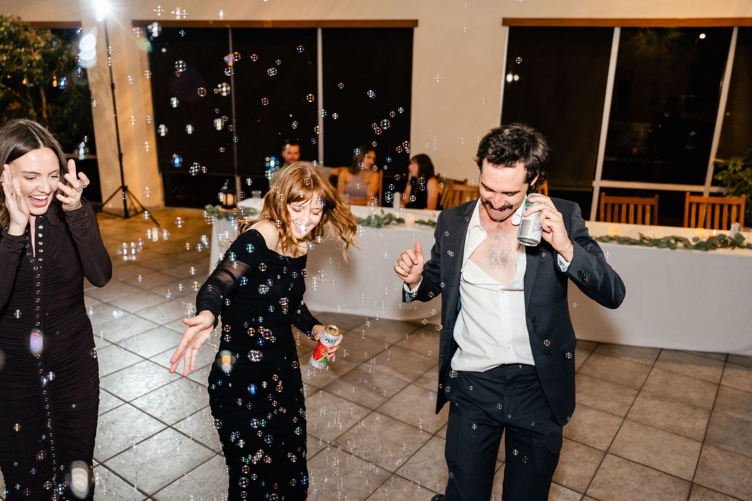 Three people dance joyfully on a tiled floor at an evening party with decorative lights in the background.