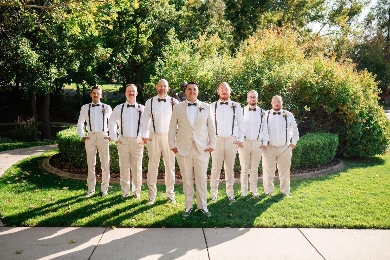 A wedding party of groomsmen wearing light beige suits and suspenders pose together on a sunny garden path.