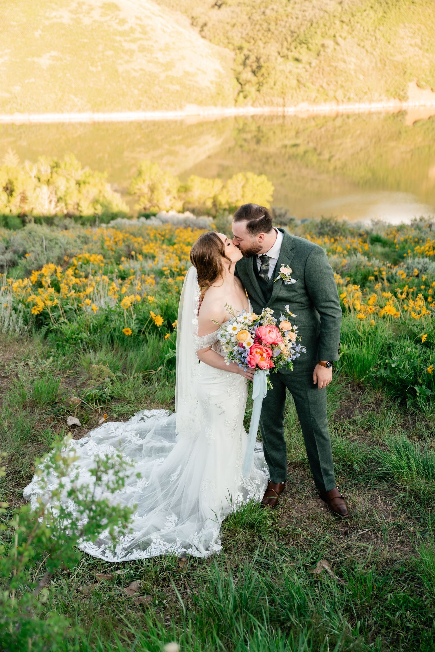 Couple shares intimate moment surrounded by yellow wildflowers with lake and mountains in background.