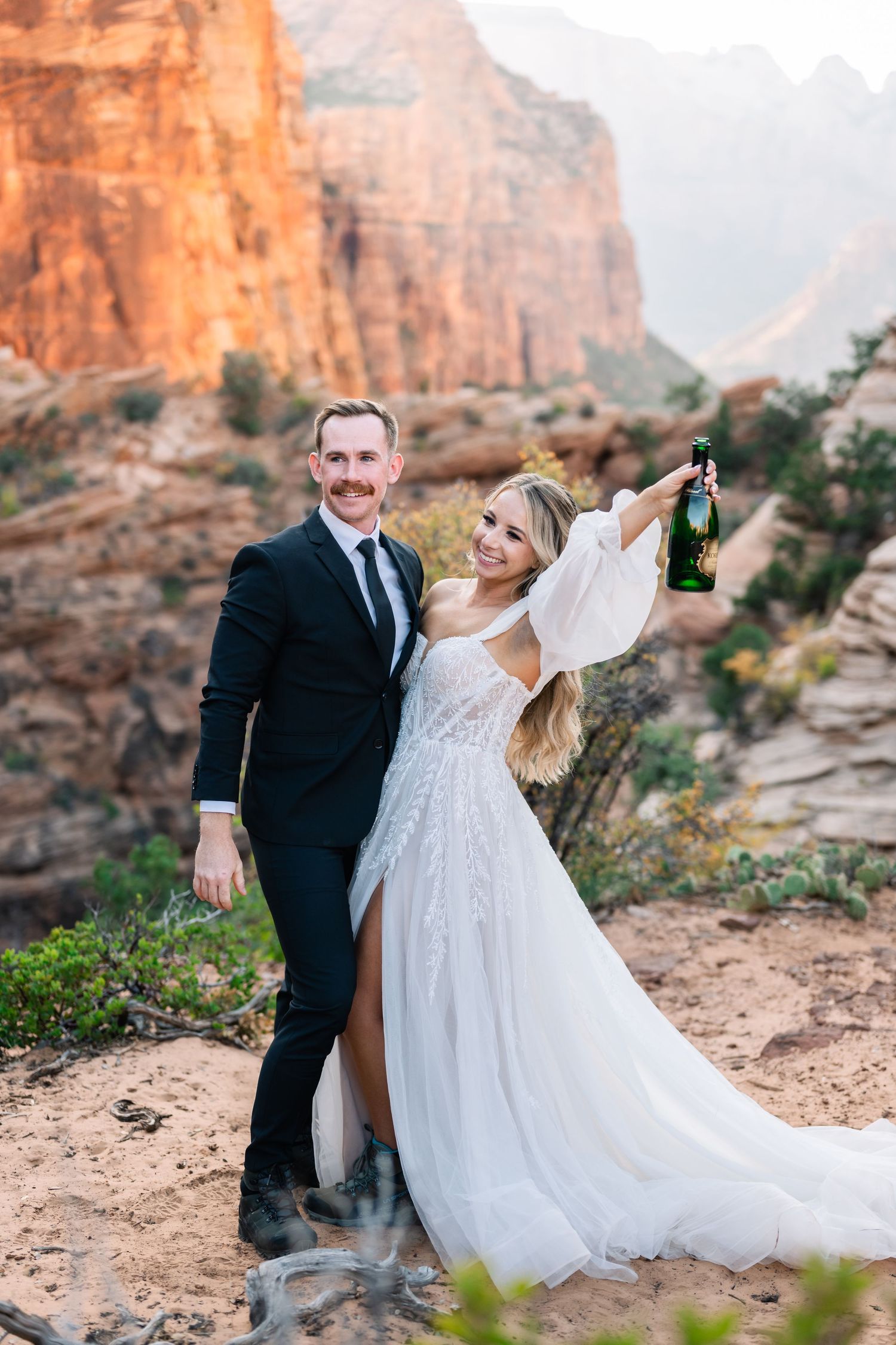 Newlyweds toast with champagne bottles against backdrop of majestic red rock formations.