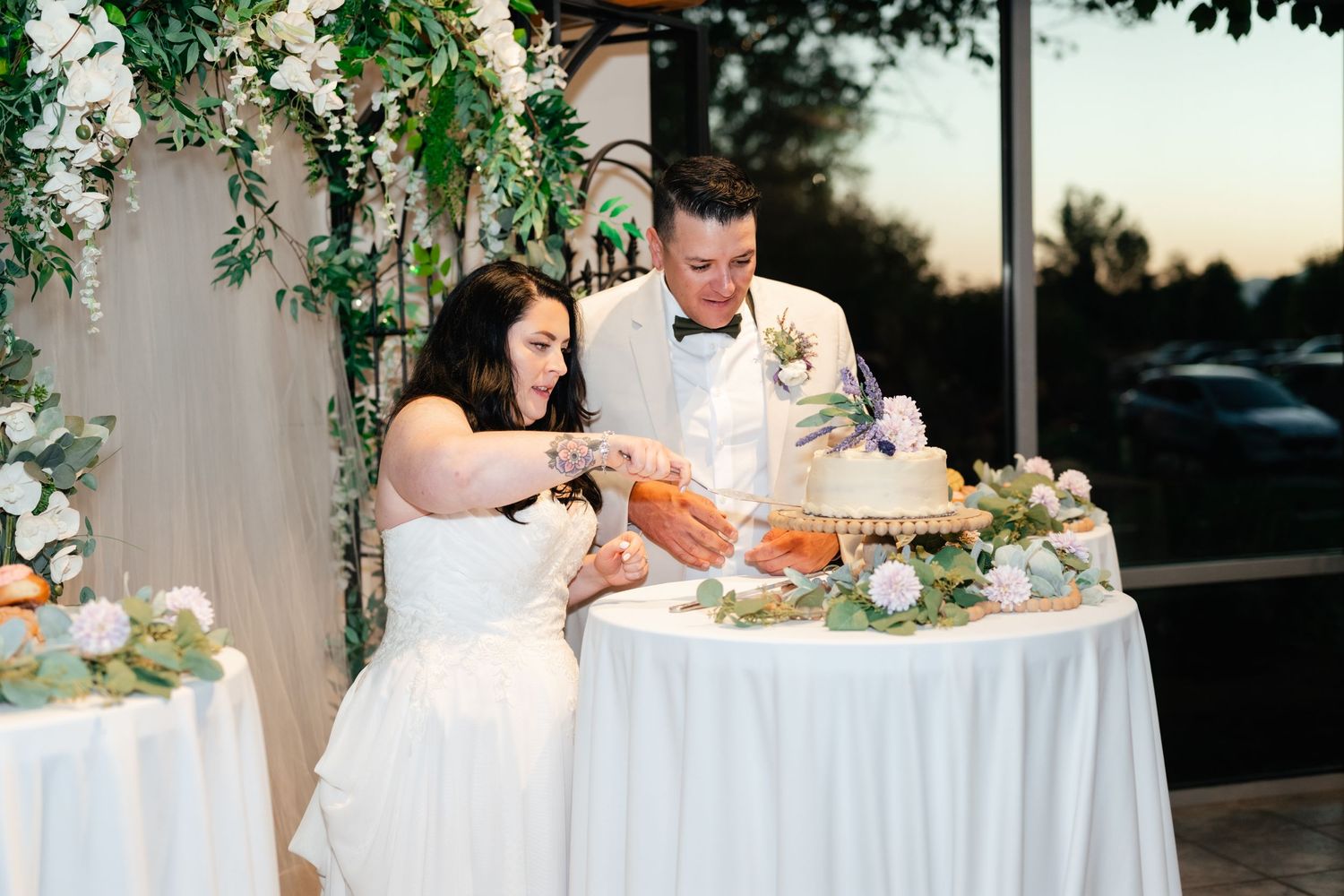 A couple cuts their wedding cake together at an outdoor reception decorated with white linens and floral arrangements.