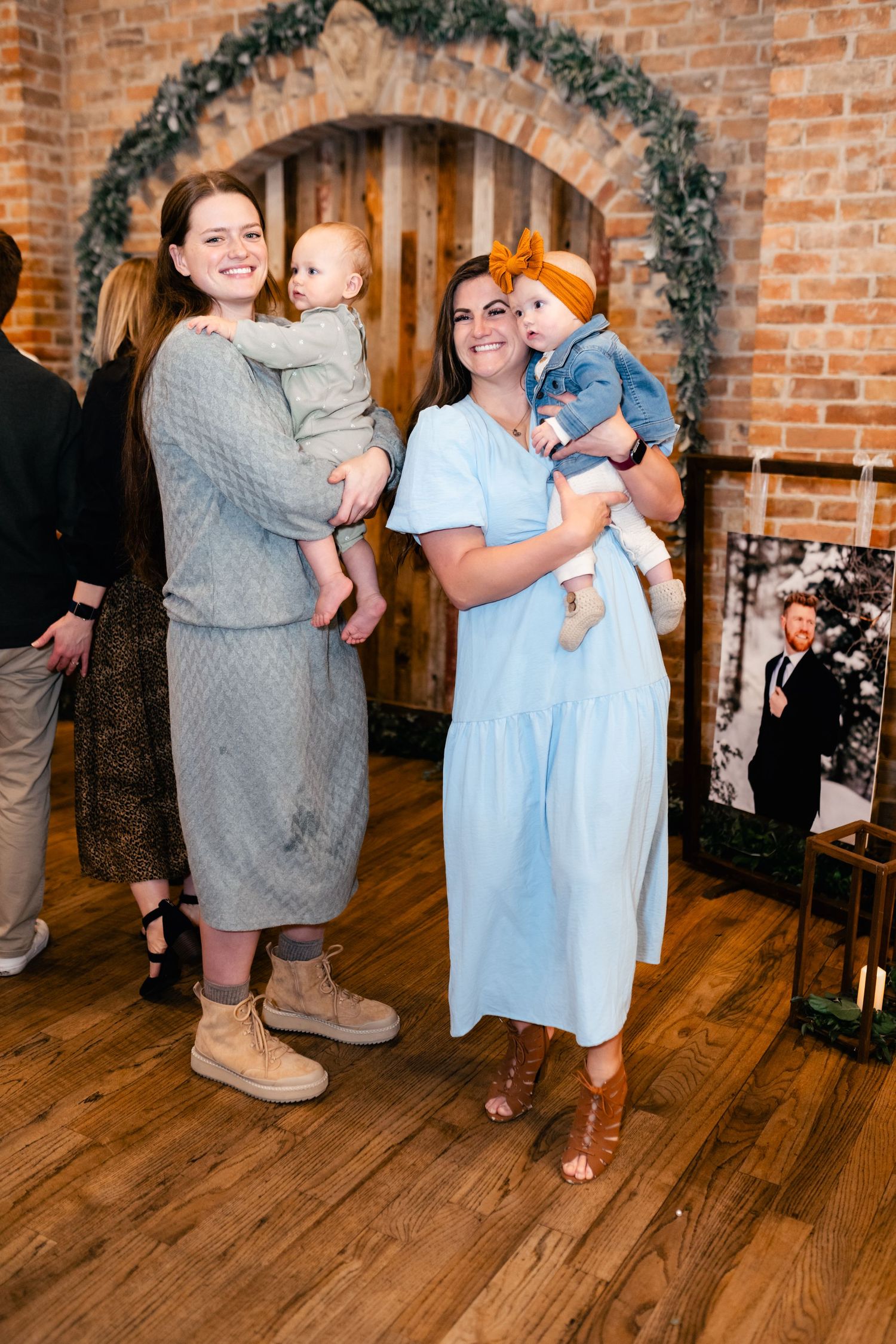 Two women hold babies while posing in front of a decorated brick archway with garland at an indoor event.