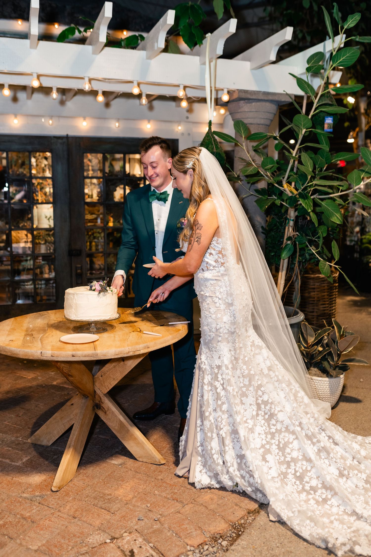 A couple cuts their wedding cake together under a pergola decorated with string lights at an outdoor wedding reception.