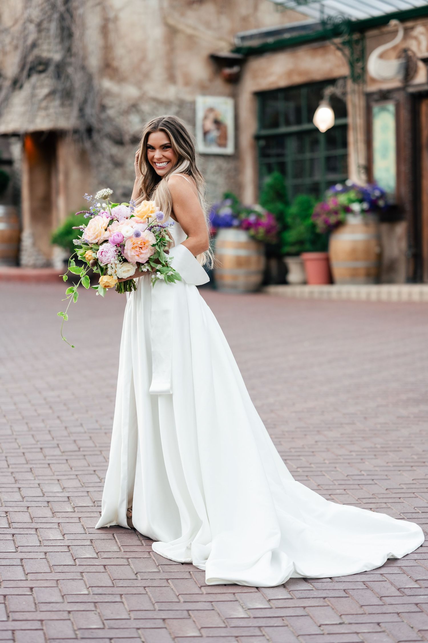 Beautiful long white wedding dress with flowing train on brick courtyard with hanging floral arrangements and rustic decor.