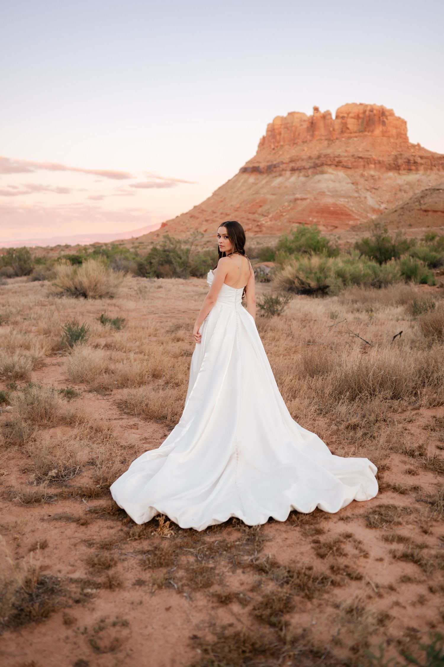 Bride in strapless white gown with train walks through desert with red rock butte at sunset.
