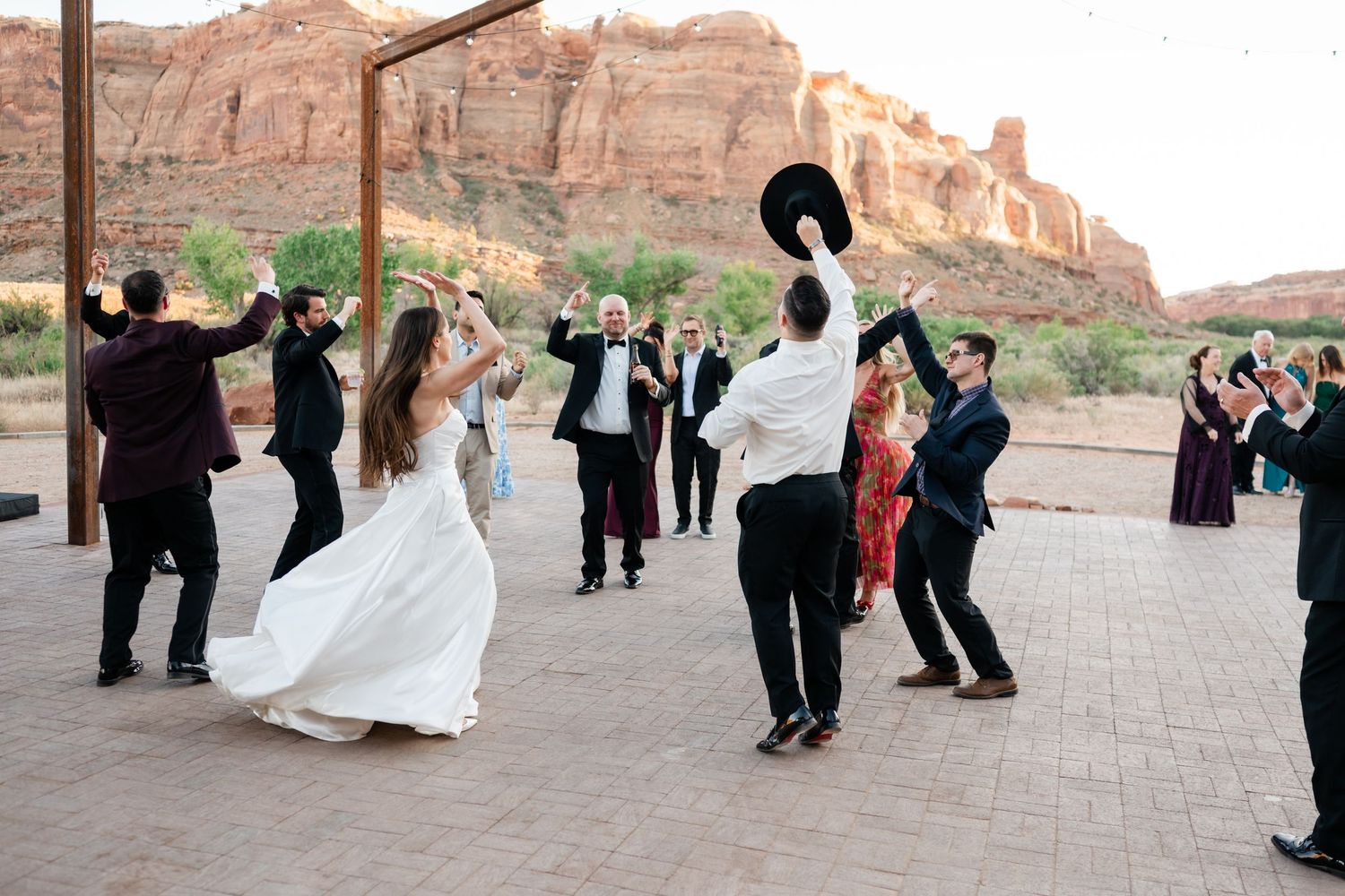 Bride and groom dance with guests at outdoor wedding reception with stunning red rock canyon backdrop in desert setting.