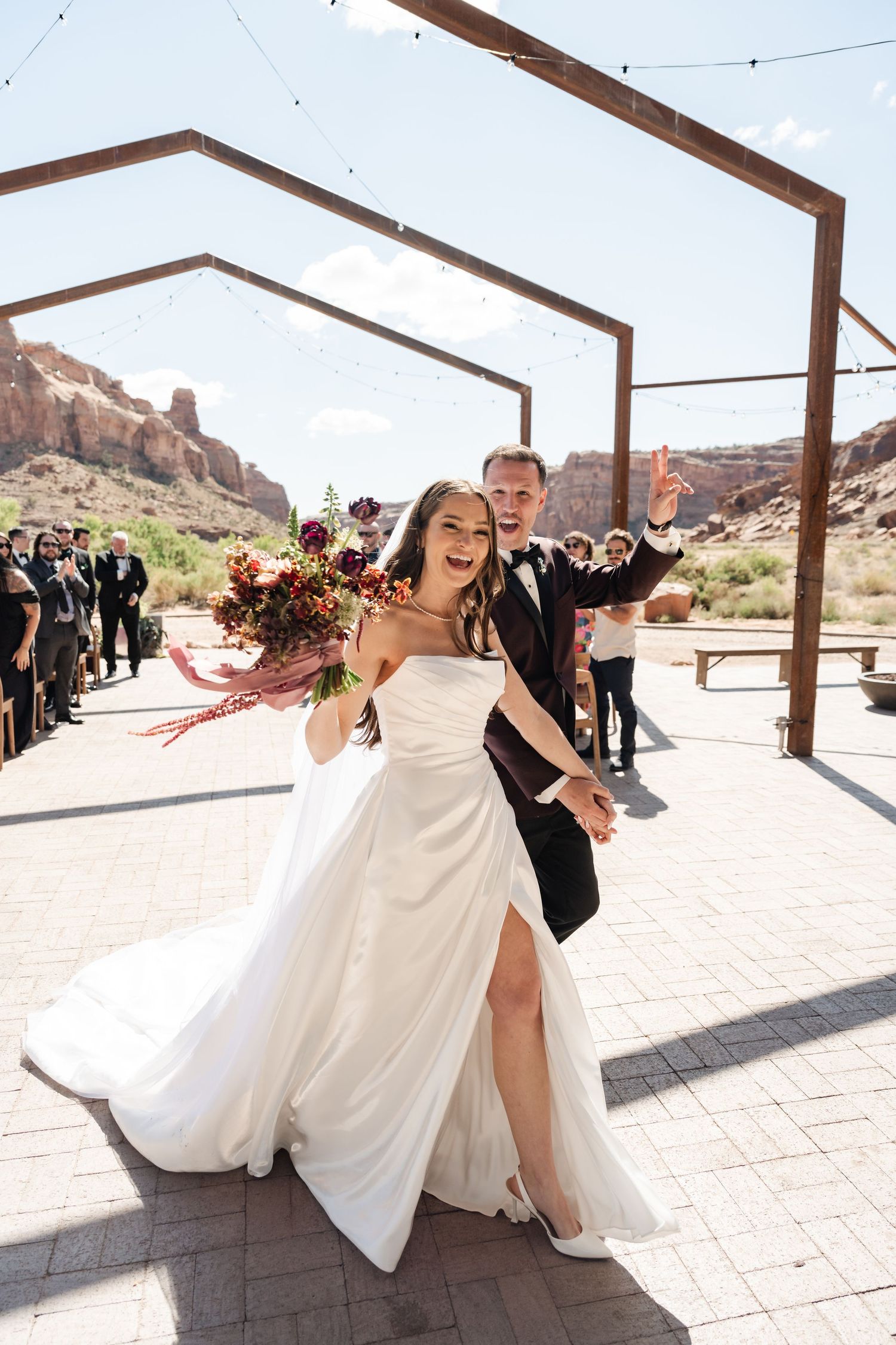 Joyful bride in white strapless gown and groom in black tuxedo celebrate after outdoor desert wedding ceremony.