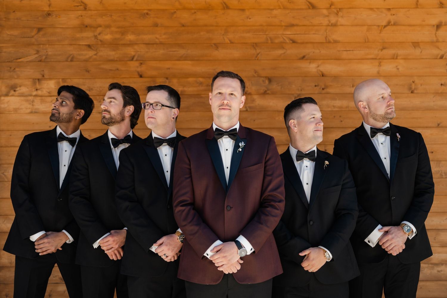 Groom in burgundy tuxedo stands with five groomsmen in black tuxedos and bow ties inside a wooden barn venue.