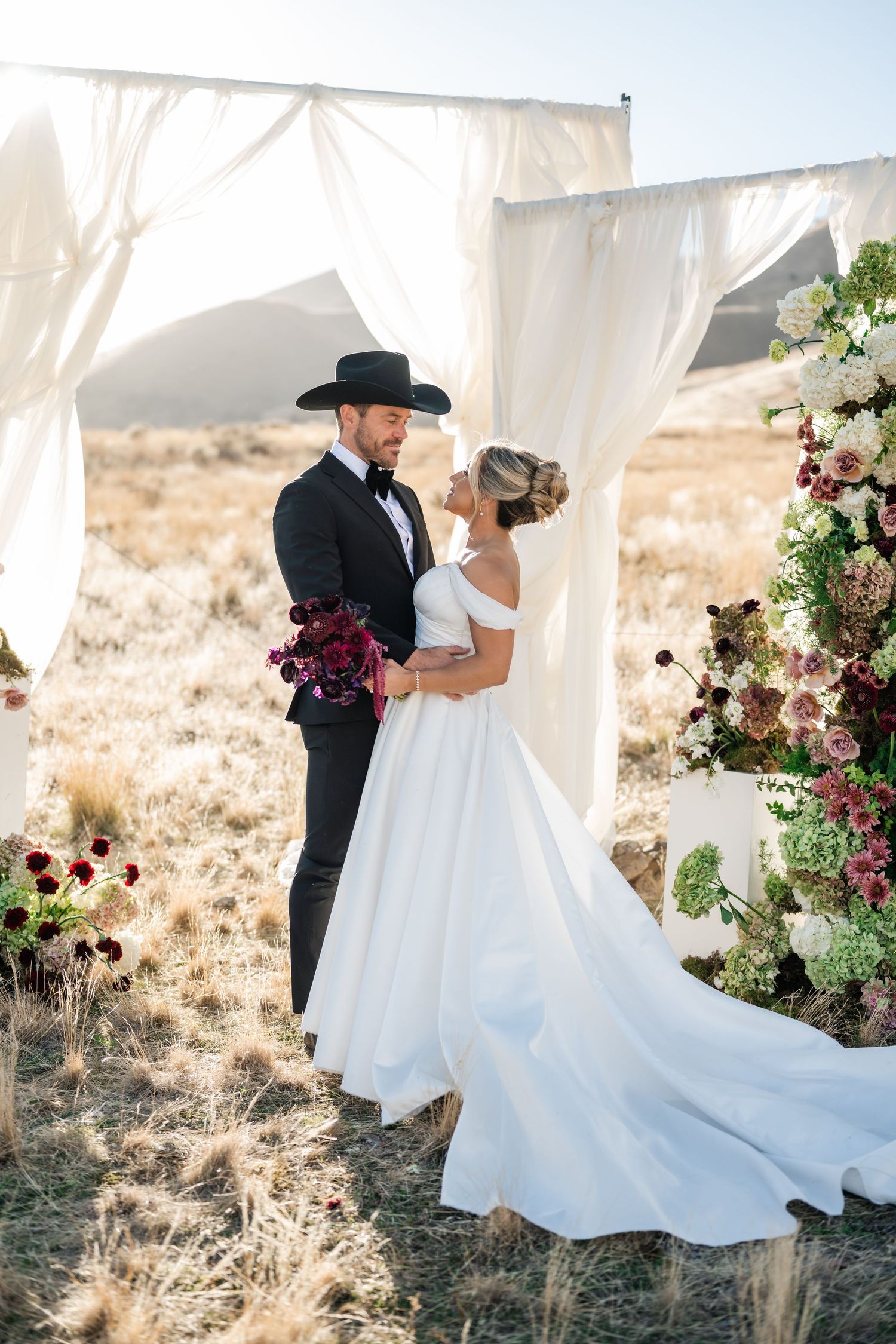 Romantic wedding couple embraces under flowing white canopy with burgundy floral arrangements in desert landscape.