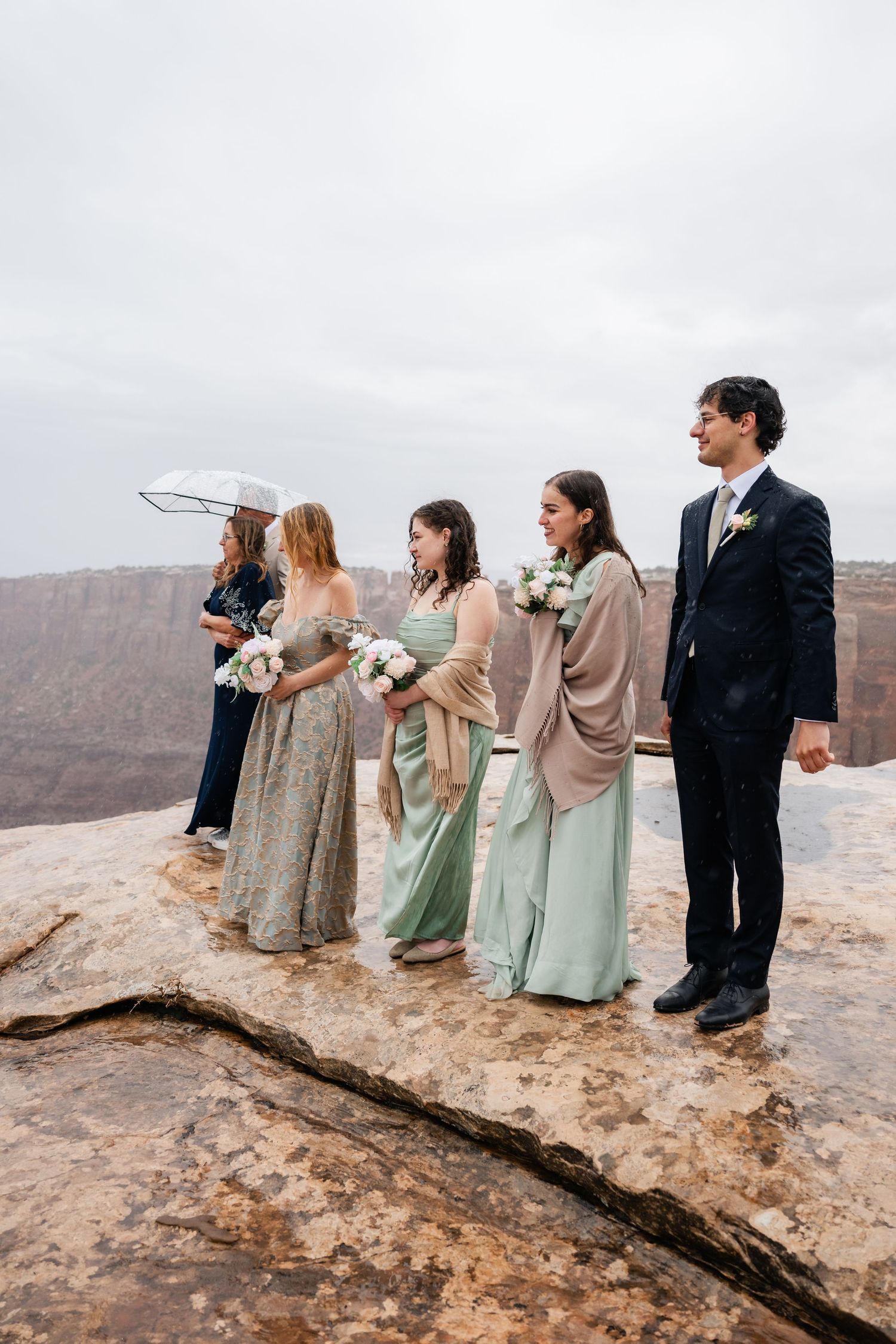 Wedding party stands in a line on a rocky cliff edge looking out over a dramatic desert canyon landscape.