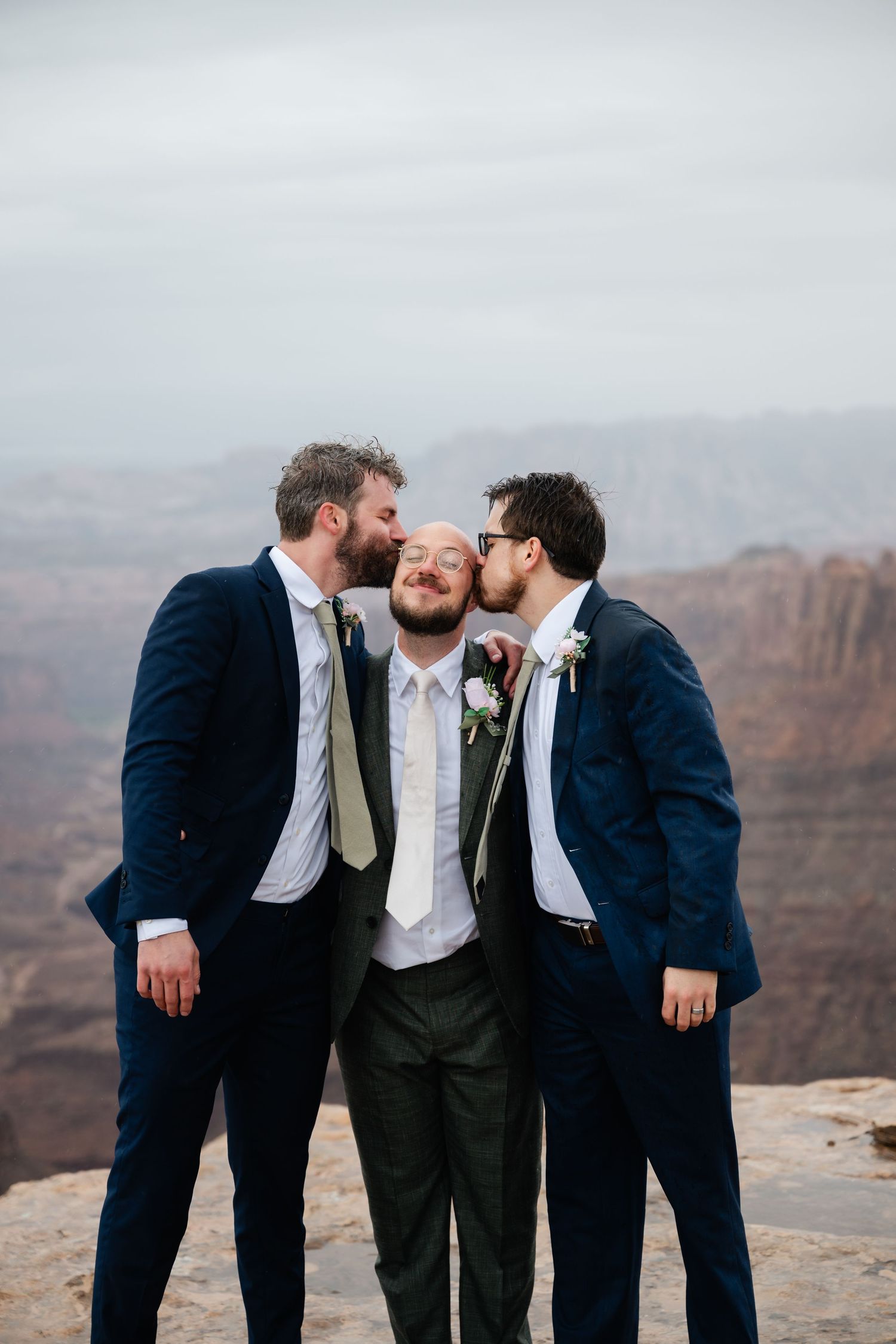 A group in suits shares a lighthearted moment at the edge of a scenic canyon overlook.