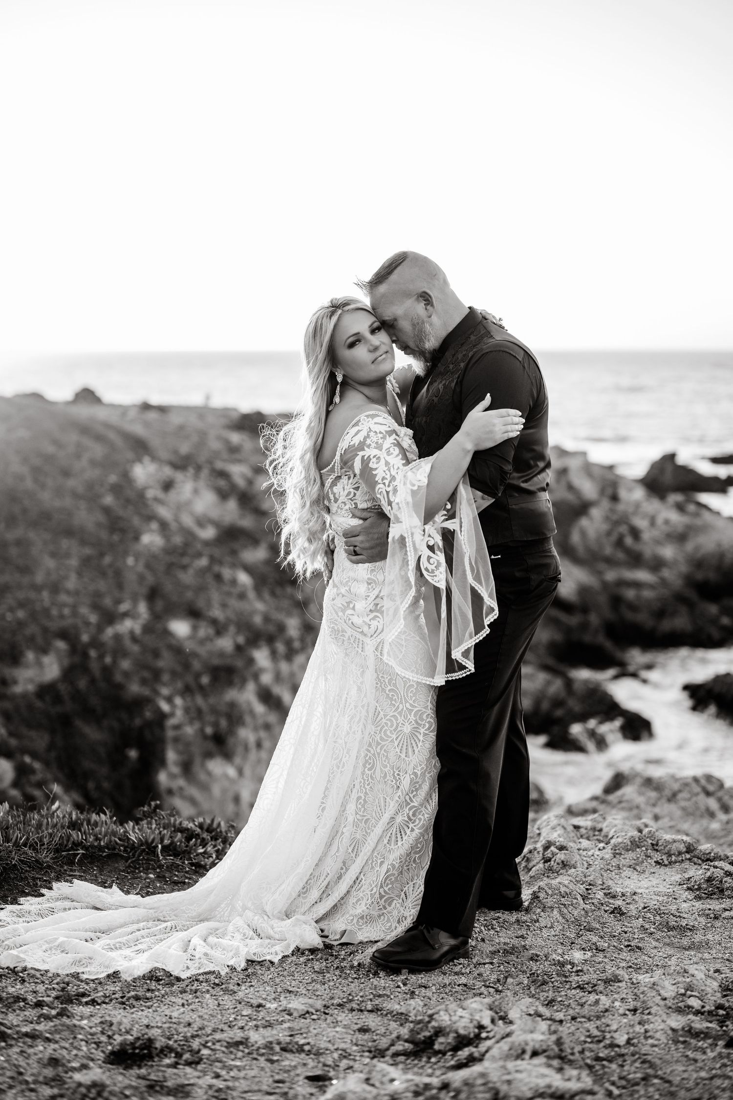 A romantic black and white wedding photoshoot on a rocky beach coastline with dramatic ocean views in the background.