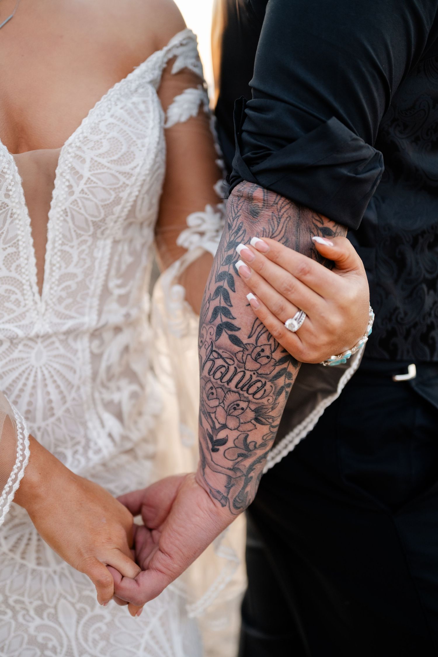Close-up of tattooed hands holding each other against white lace wedding dress.