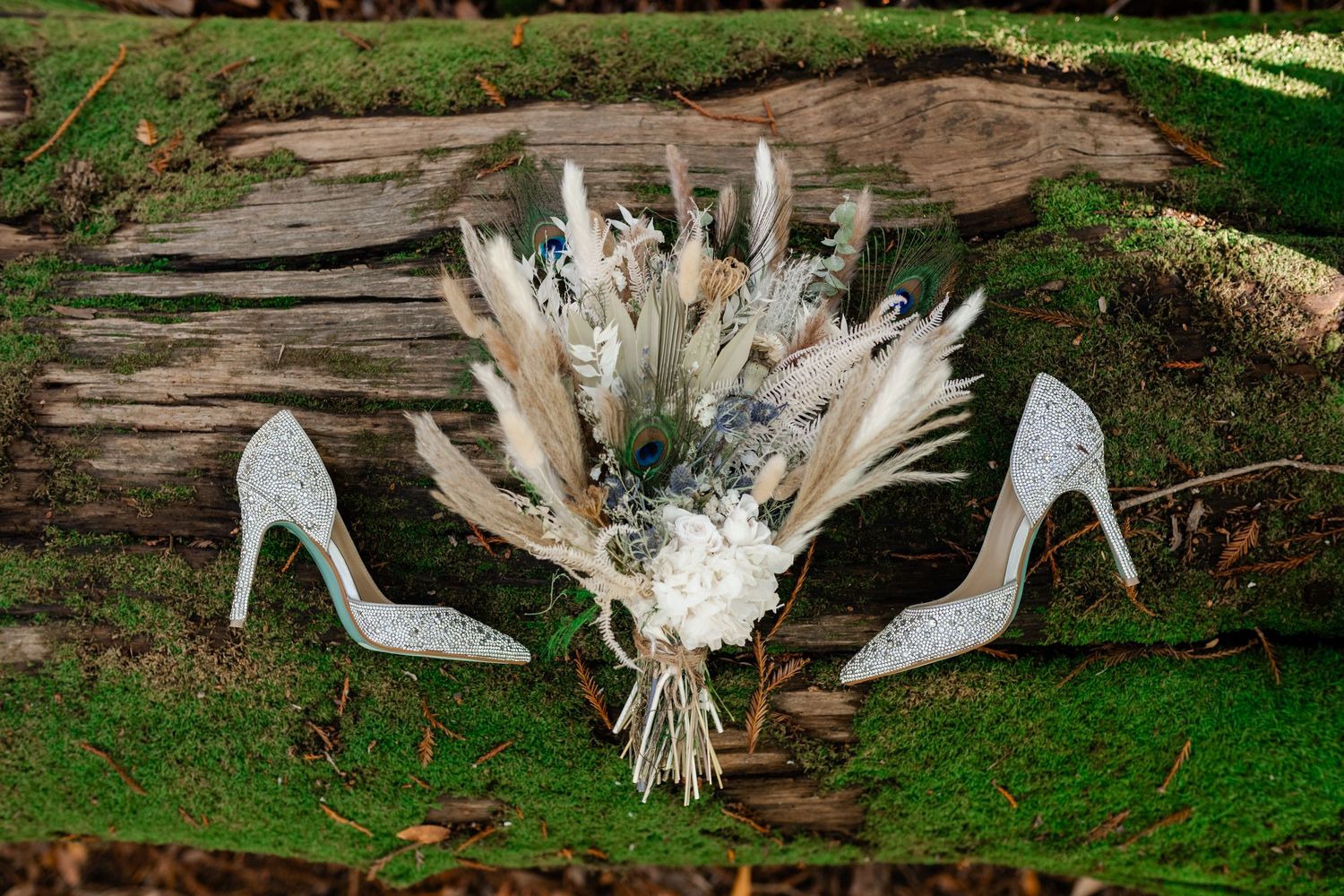 White bridal shoes adorned with delicate feathers and pearls sit elegantly on rustic wooden logs in a garden setting.