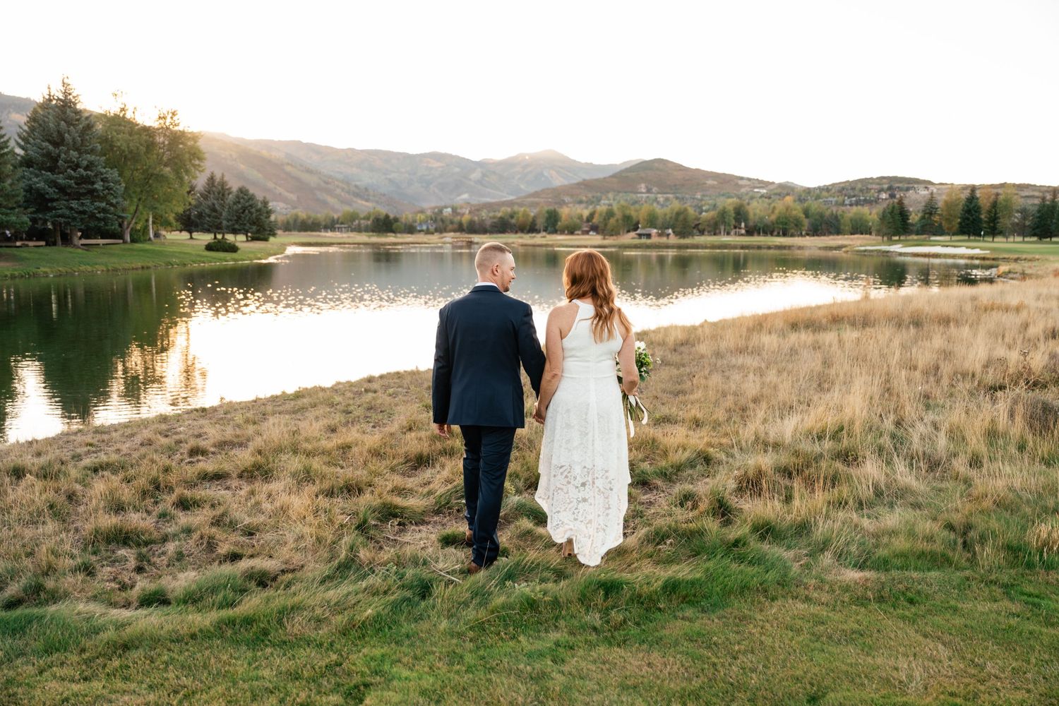 A couple walks hand in hand along a serene lakeside with mountains and golden grass in the background.