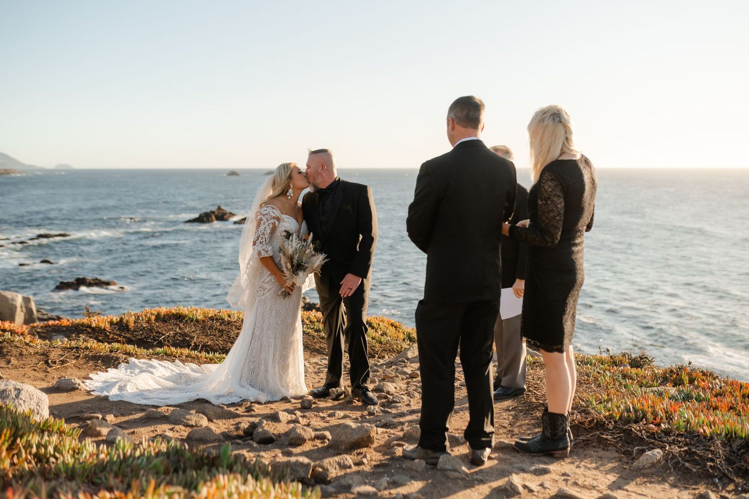 A small wedding gathering takes place on a dramatic coastal overlook with crashing waves below.
