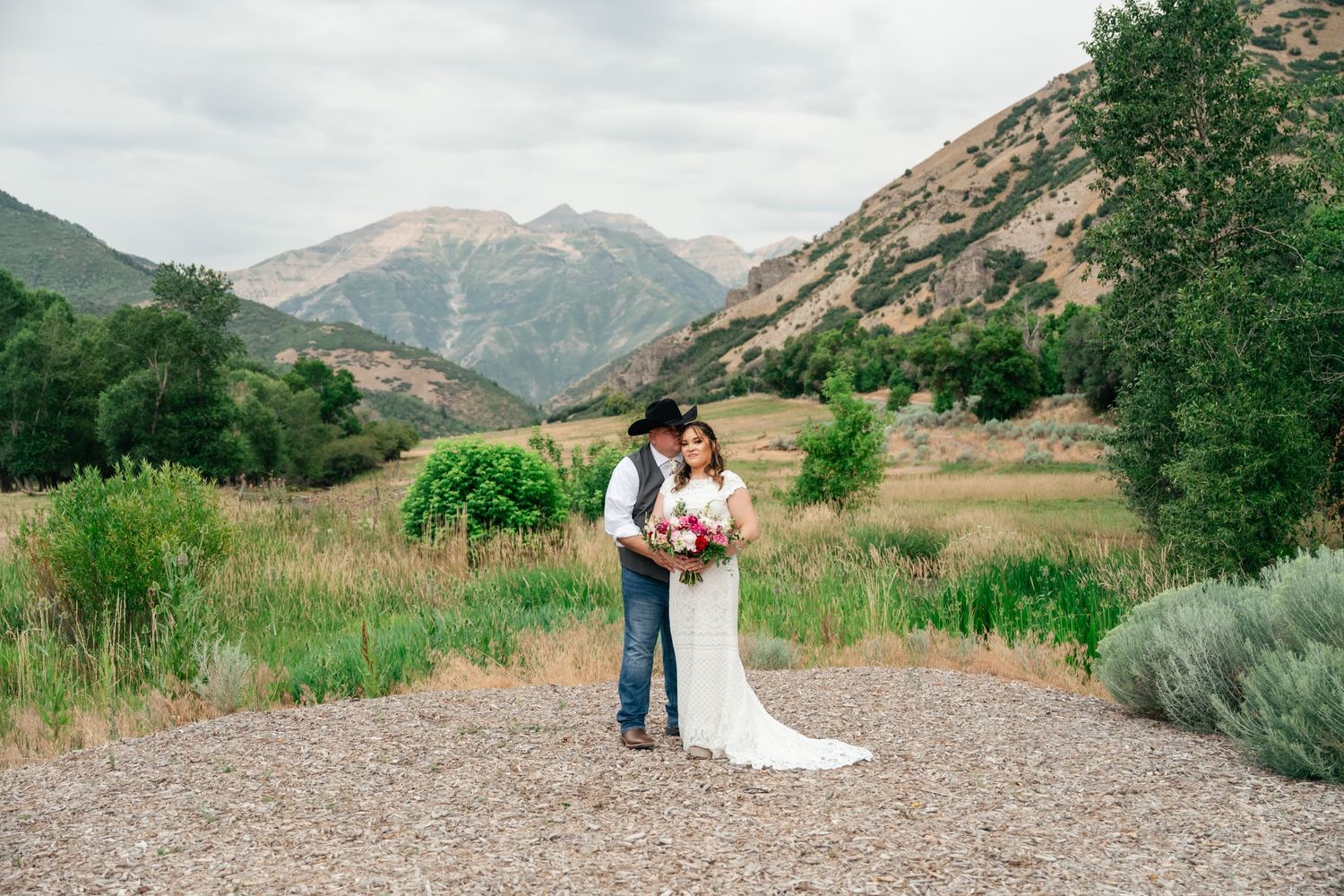 A couple in wedding attire embrace on a dirt path with majestic mountain peaks and lush greenery in the background. Conrad Ranch.
