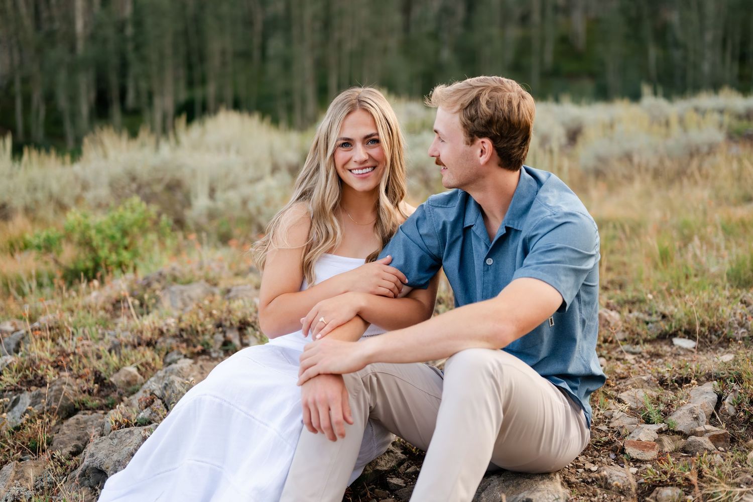 Couple sitting on rocks in a meadow, woman in white dress smiling, man in blue shirt, with trees in background.