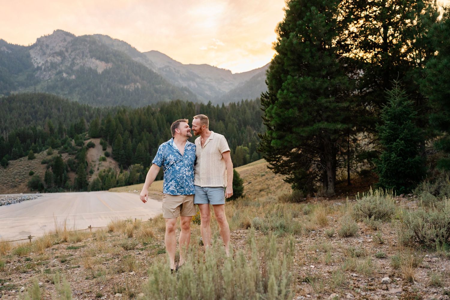 LGBTQ friendly photographer. Two men share a romantic moment on a mountain hillside at sunset with pine trees and peaks in the background.