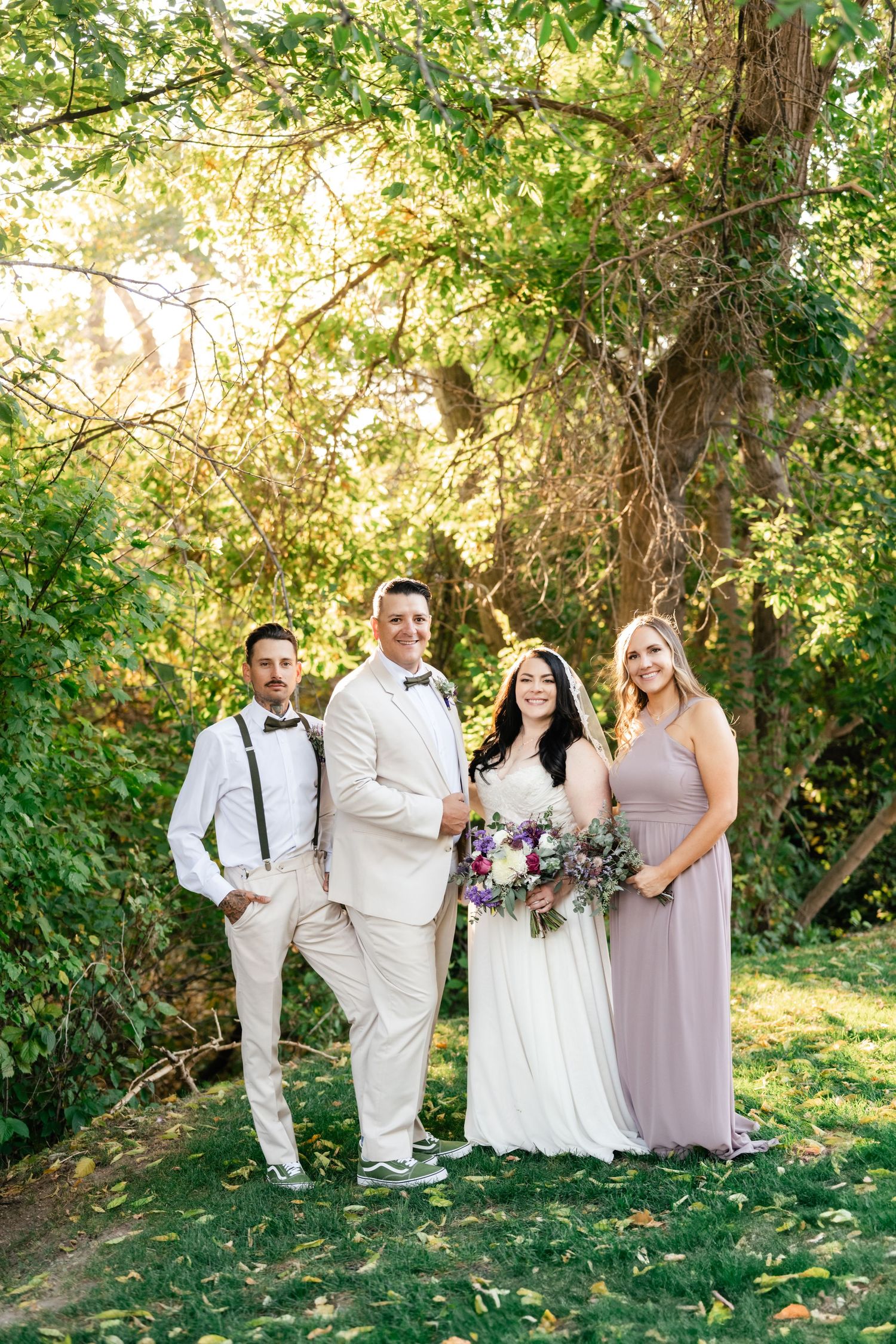 Wedding party in formal attire poses outdoors in a sunlit garden setting with greenery and trees in background.
