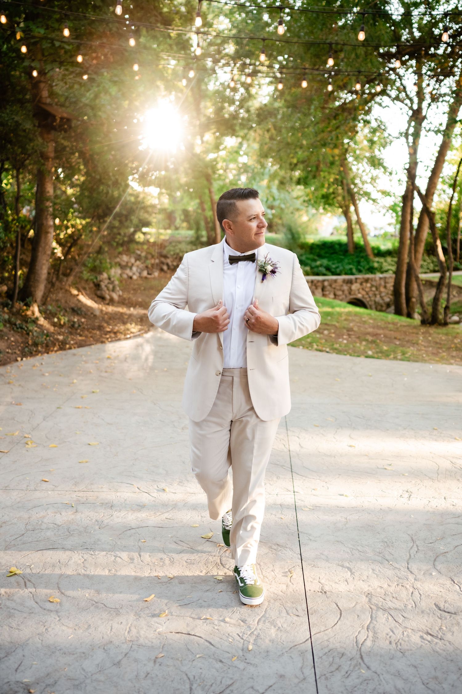 A groom in a light-colored suit and bowtie poses on a sunlit garden path surrounded by trees.
