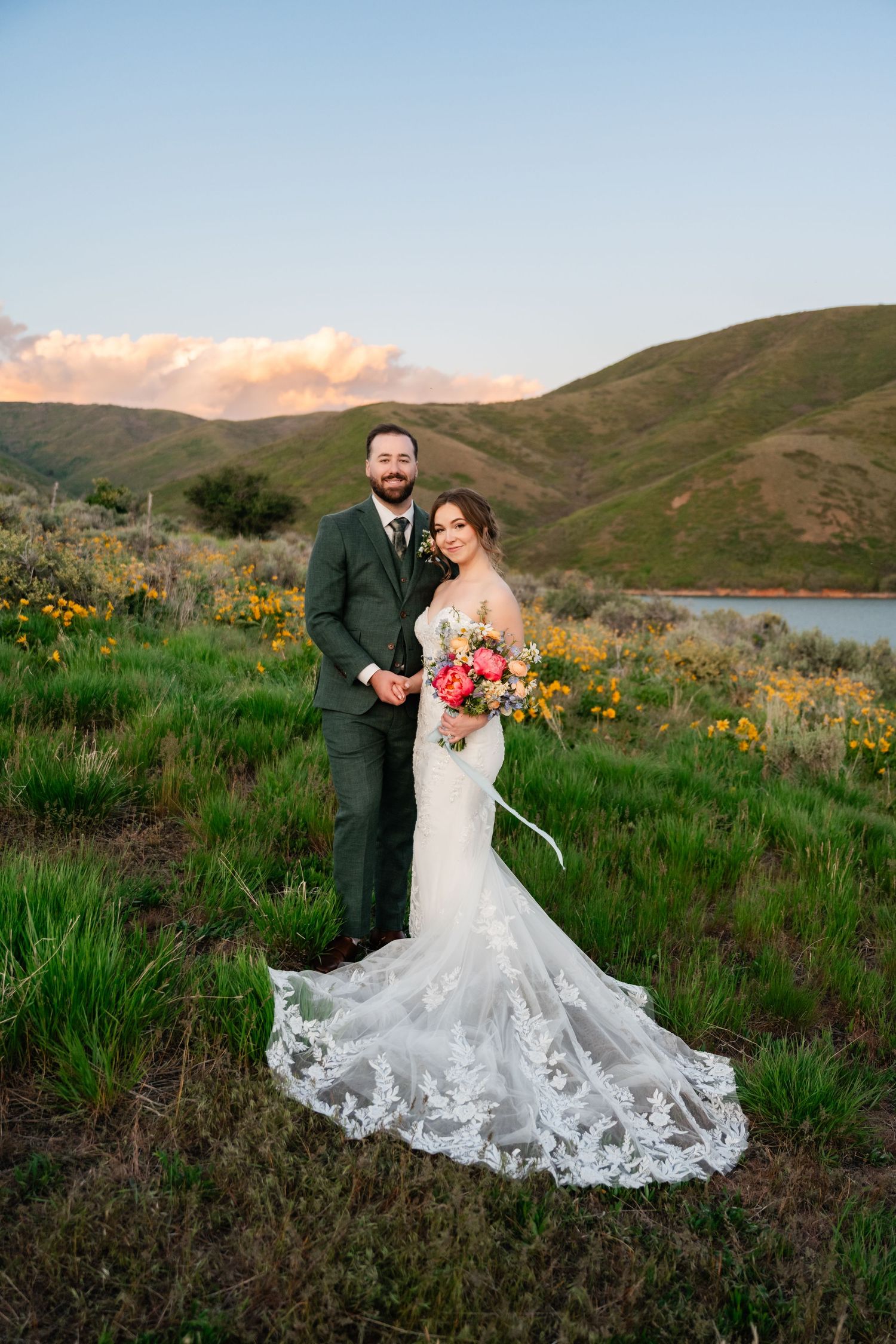 Newlyweds pose in mountain landscape with bride's lace train and colorful wildflowers.