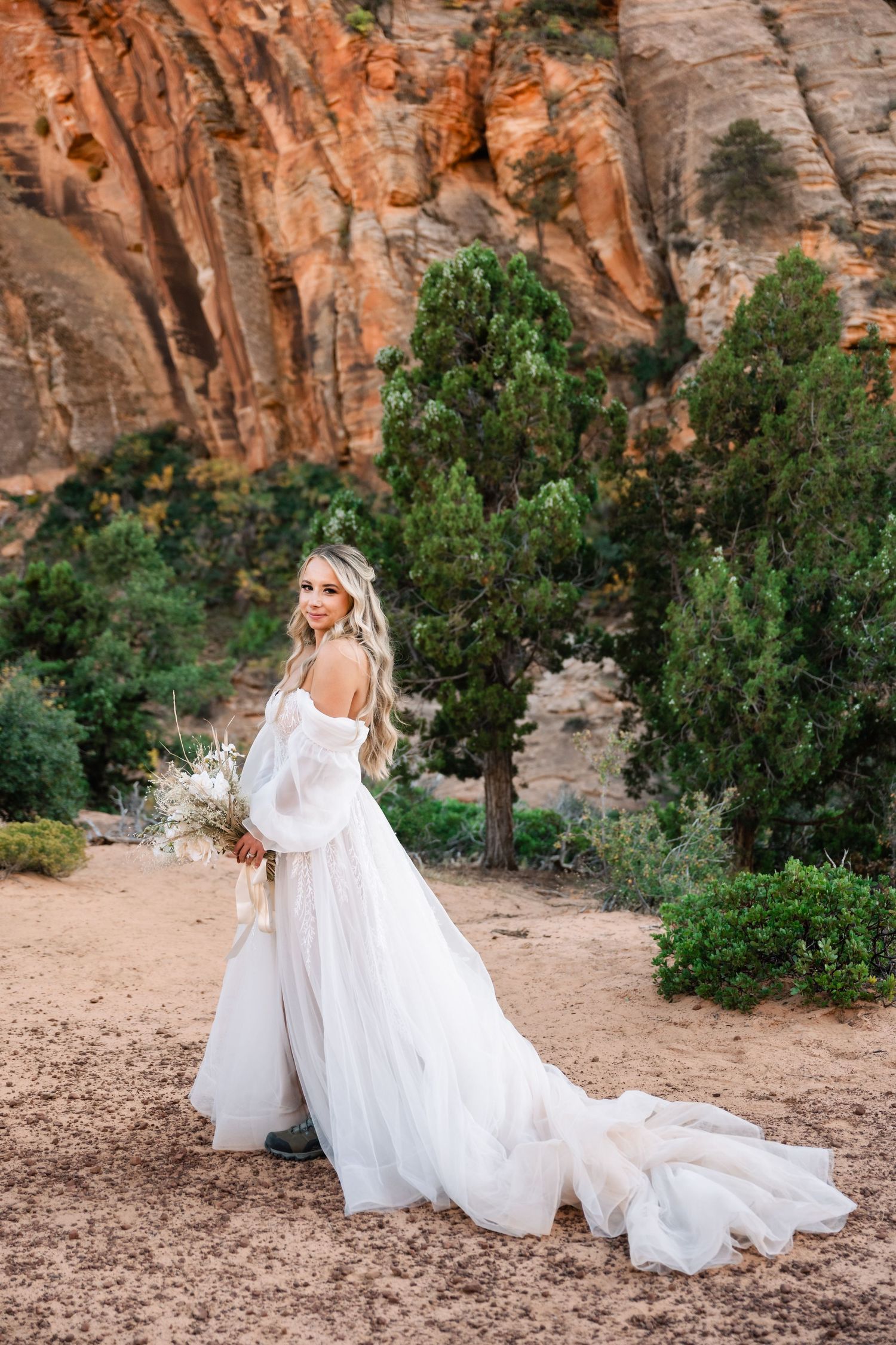 A bride in off-shoulder gown poses gracefully against majestic desert canyon backdrop.