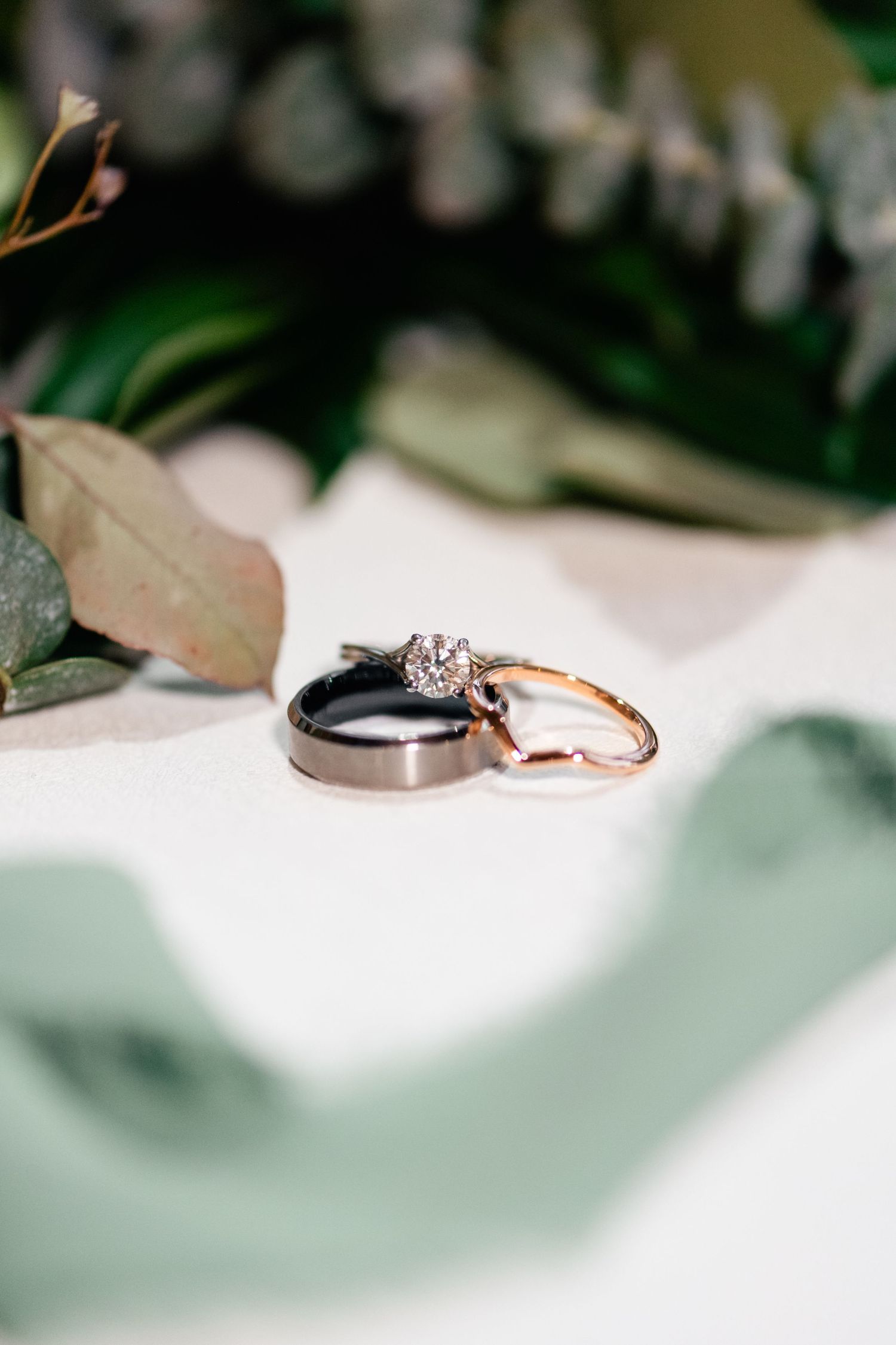 Wedding rings photographed on white surface with soft green foliage in background, featuring rose gold and grey metal bands.