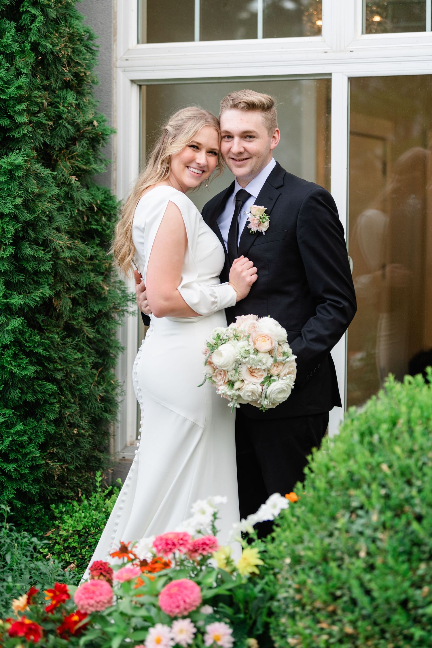 Newlywed couple poses together outdoors with pink flowers and greenery. Sleepy Ridge Wedding Photographer.