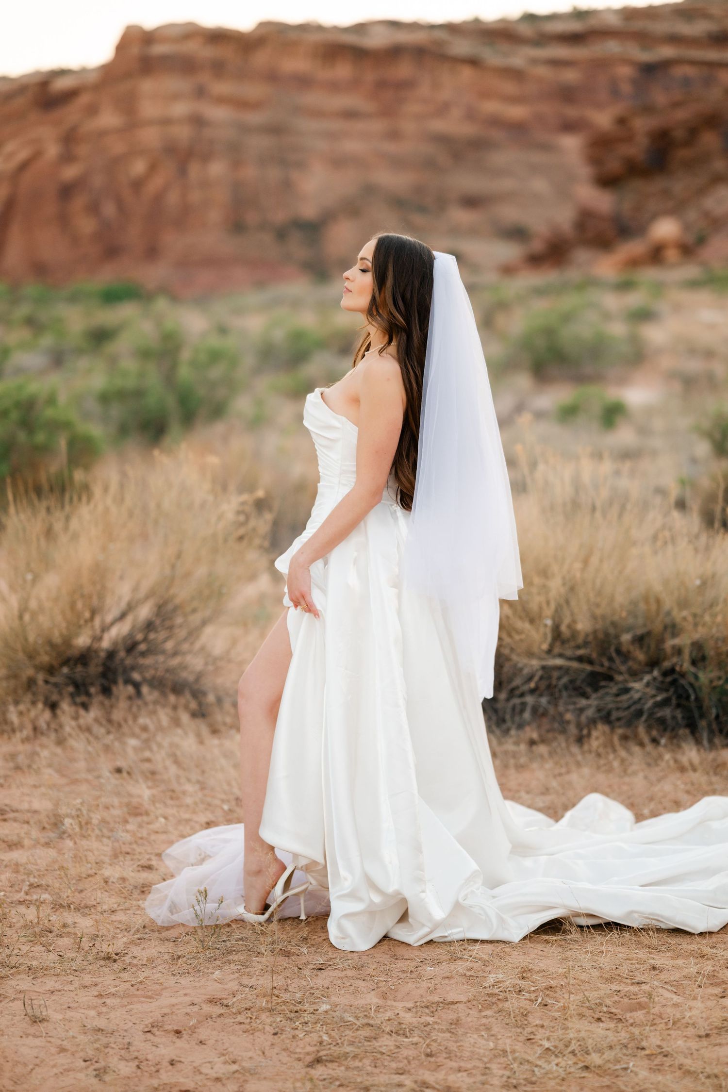 Bride in strapless white wedding gown with veil poses sideways in arid desert setting.