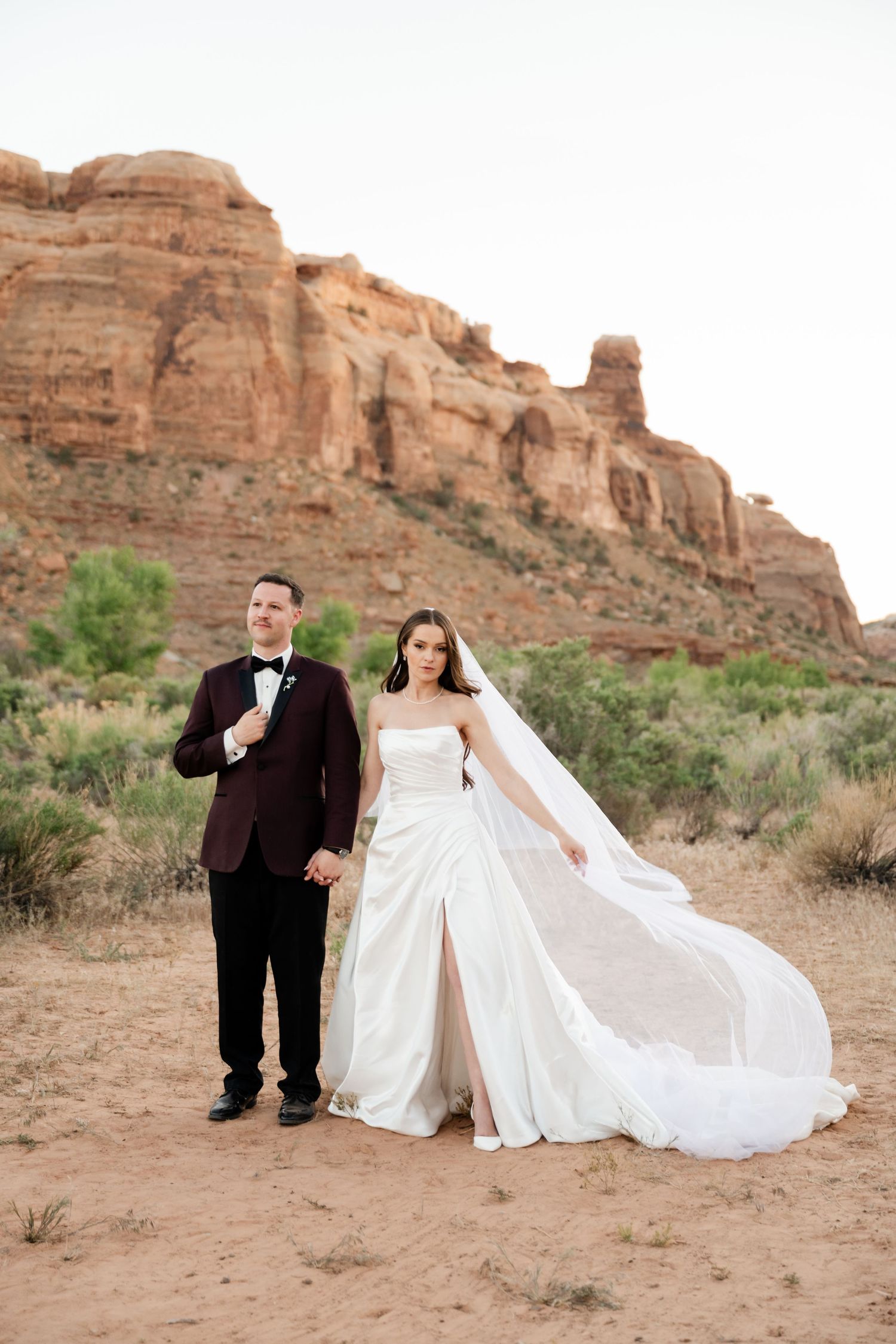 Bride in flowing white gown with veil and groom in burgundy tuxedo pose before dramatic red rock canyon walls.