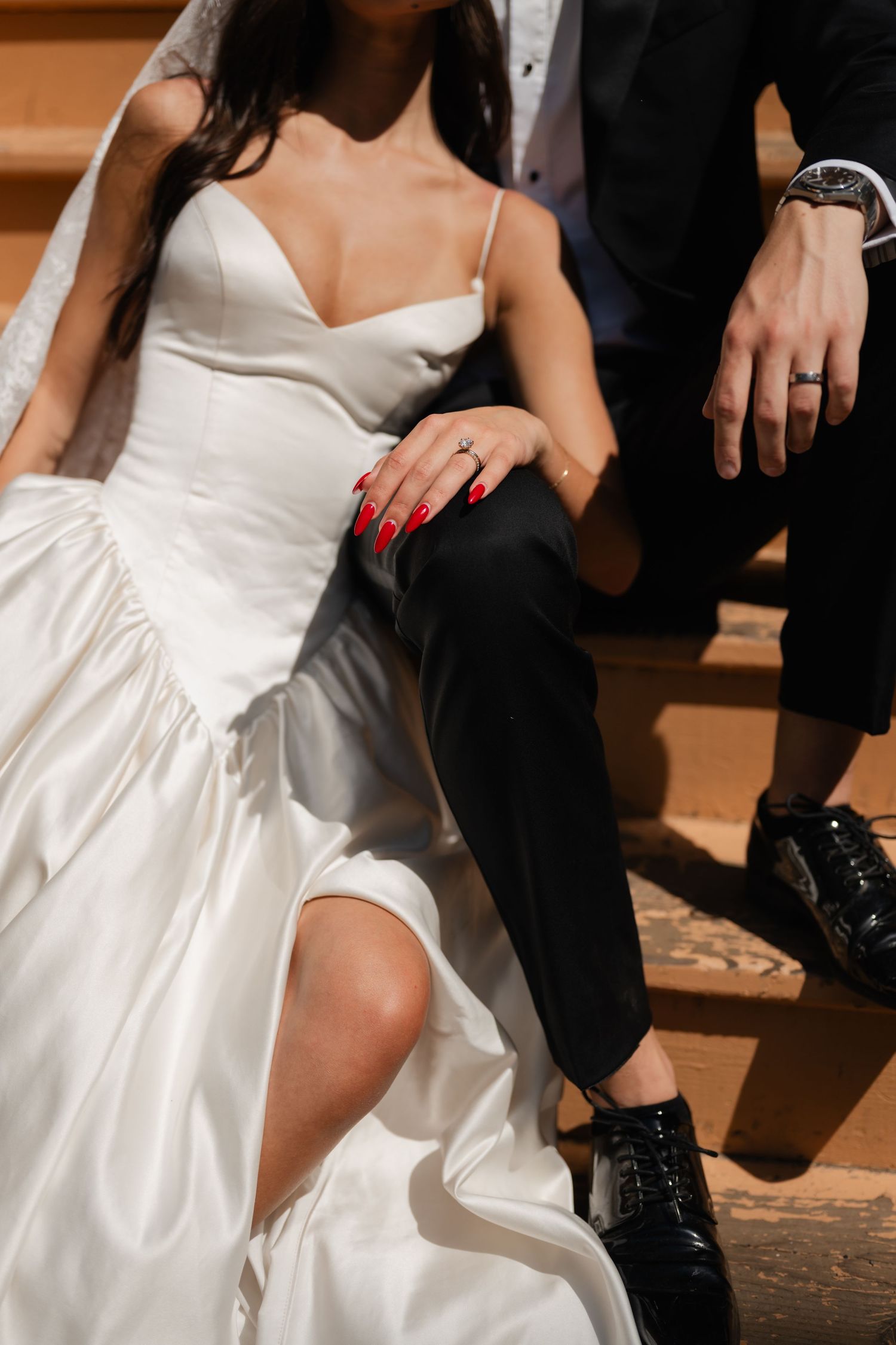 Bride in white satin gown sits beside groom in black suit on wooden steps, her red nails resting on his knee.