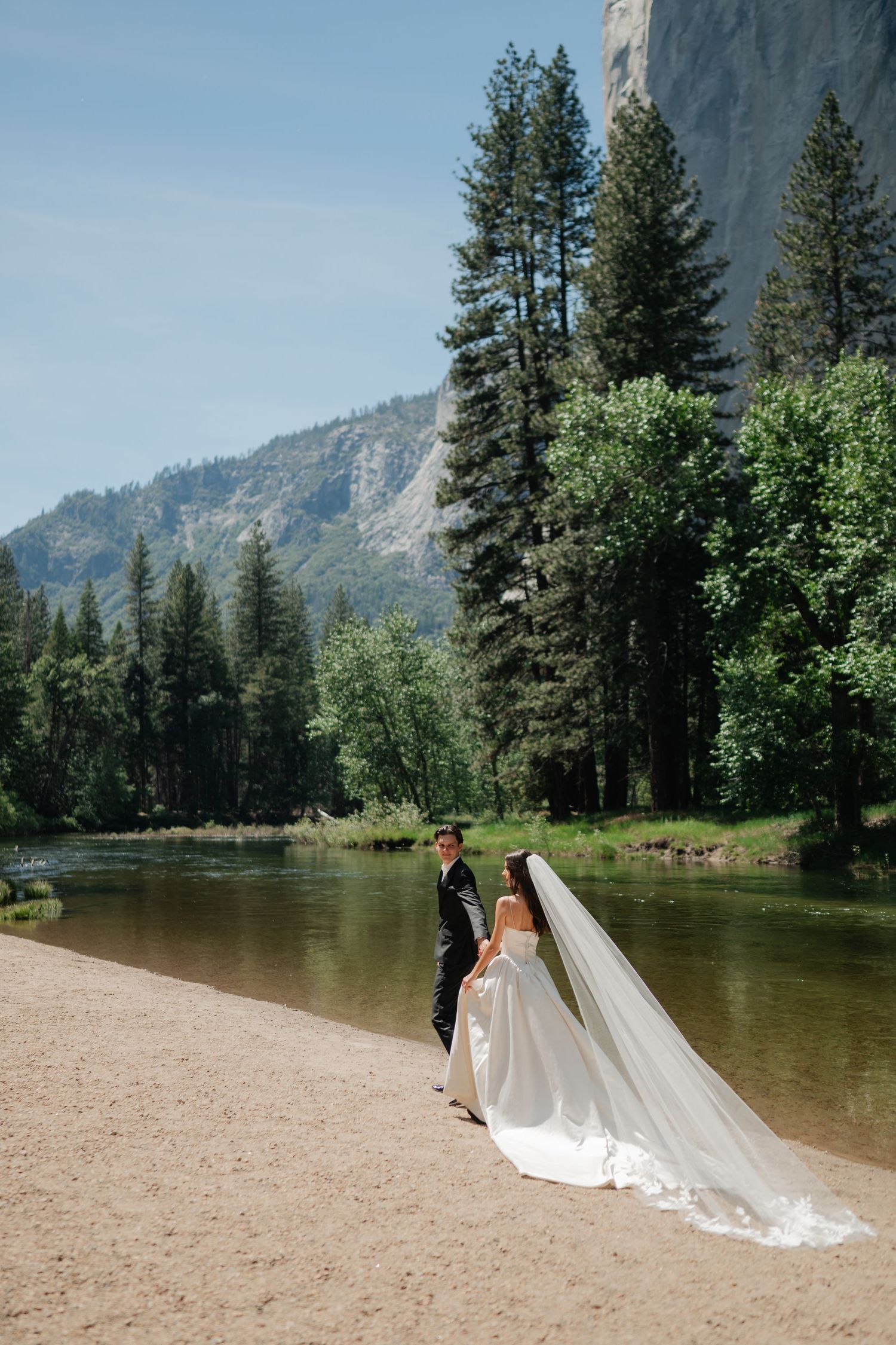 Bride and groom hold hands by a river in Yosemite, surrounded by tall pines and granite cliffs on their wedding day.