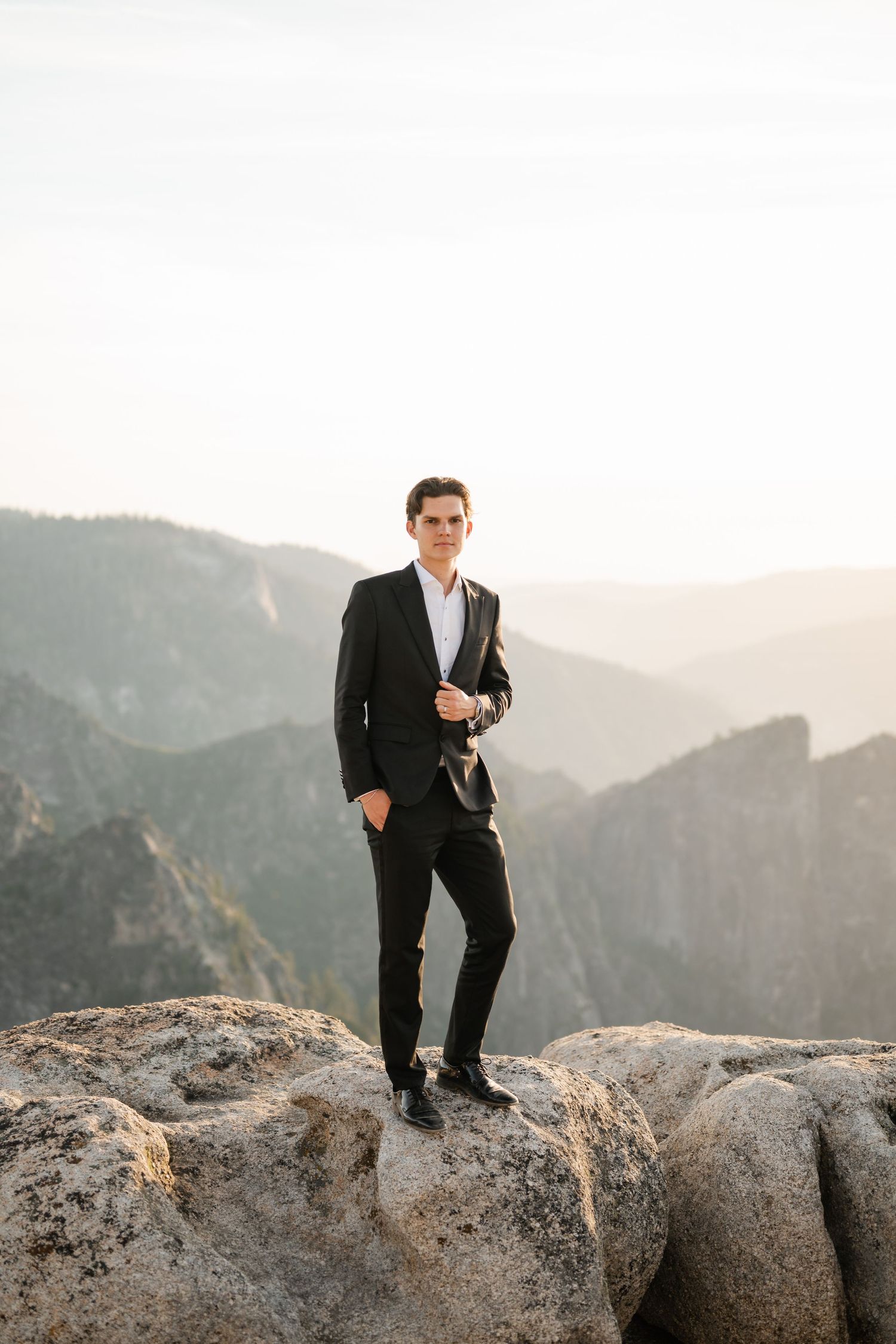 Groom in black suit stands confidently on rocky mountain overlook with misty valley backdrop.