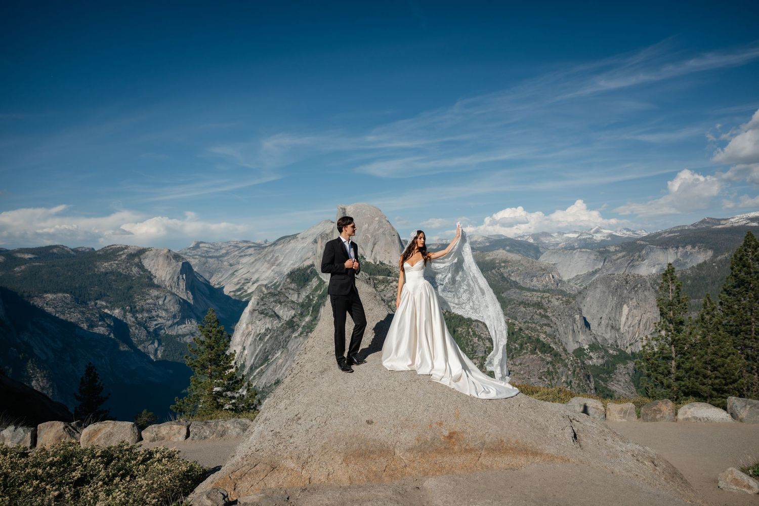 Bride and groom stand on rocky summit at Yosemite with Half Dome backdrop, wedding veil flowing in wind.