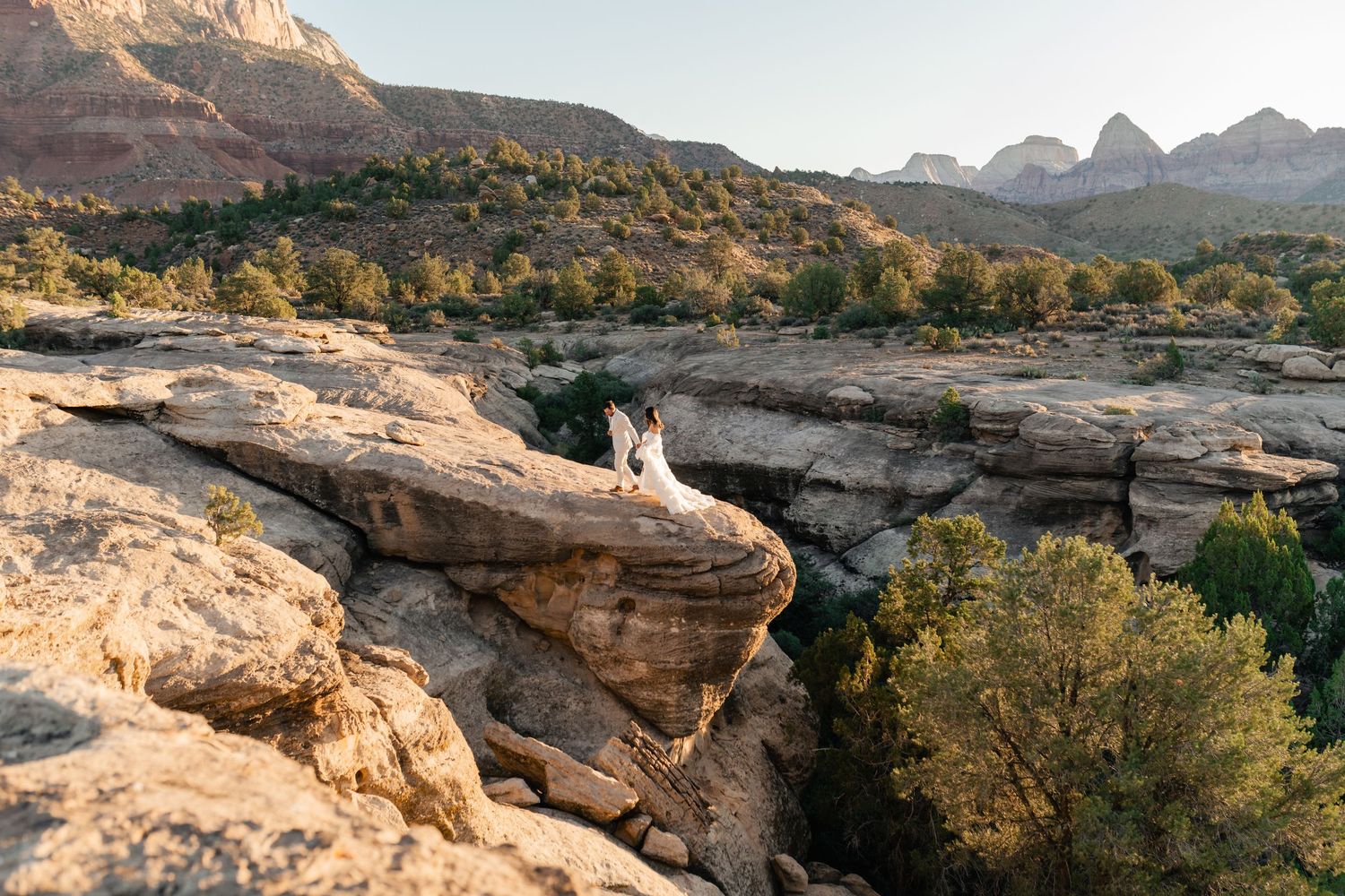 Zion Adventure Session. Wedding couple stands on dramatic red rock cliffs overlooking a vast desert canyon landscape at sunset.