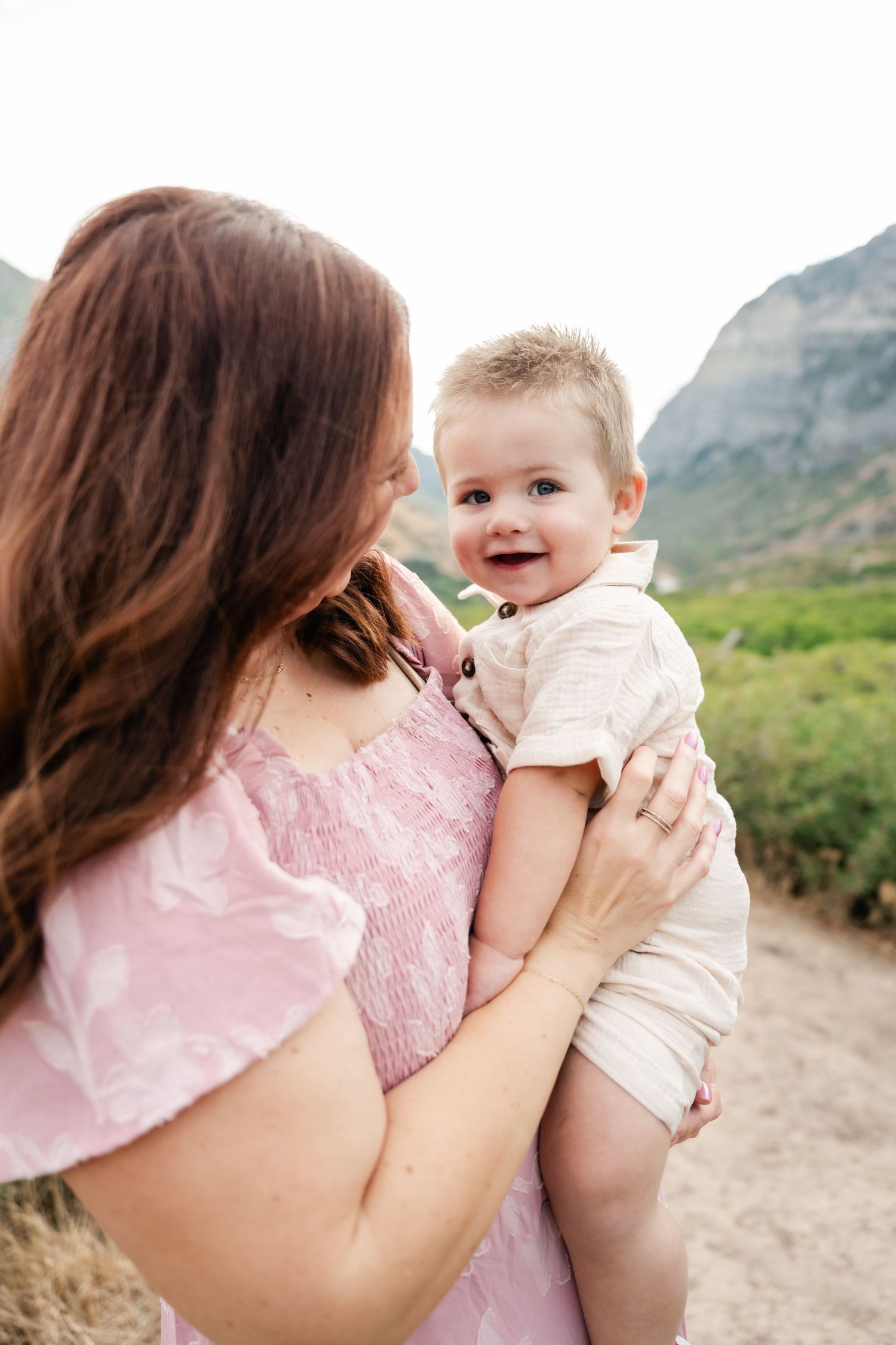 A parent in pink holds and plays with a laughing baby outdoors against a mountain backdrop in a series of tender moments.