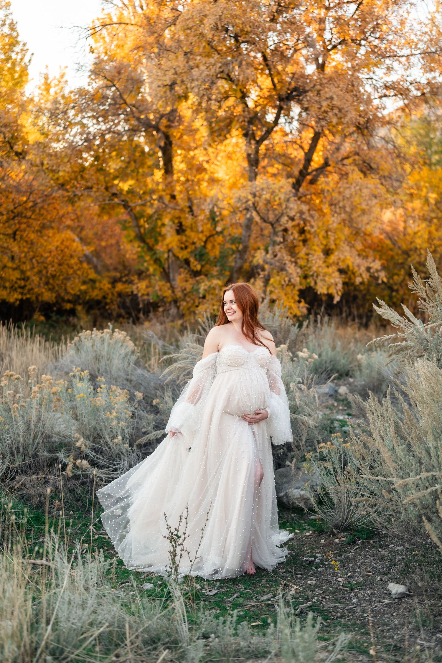 Woman in flowing white dress twirls gracefully among autumn foliage and sage brush at golden hour.