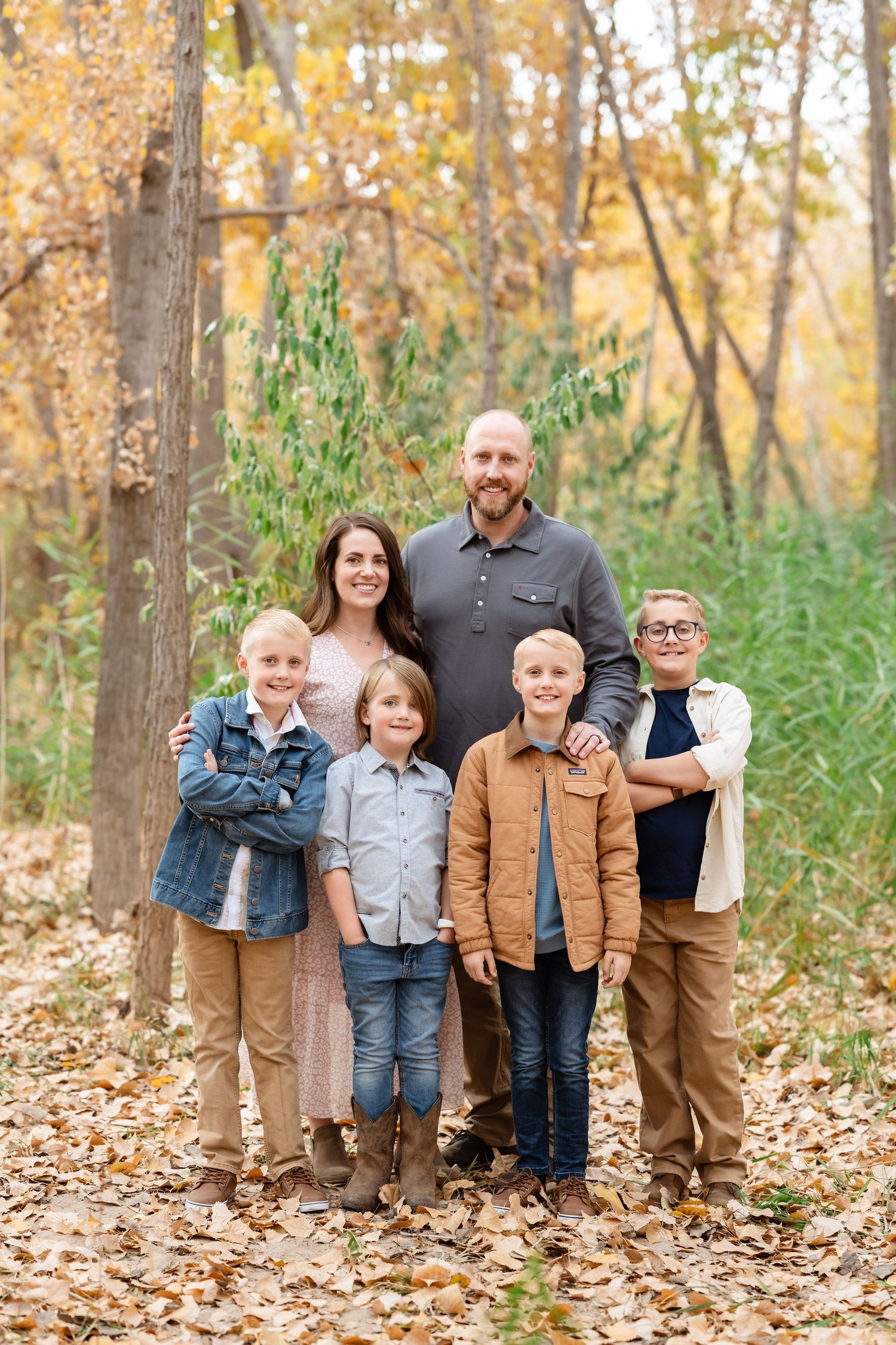 A family wearing coordinated blue and brown outfits poses together in a wooded area with golden autumn leaves.