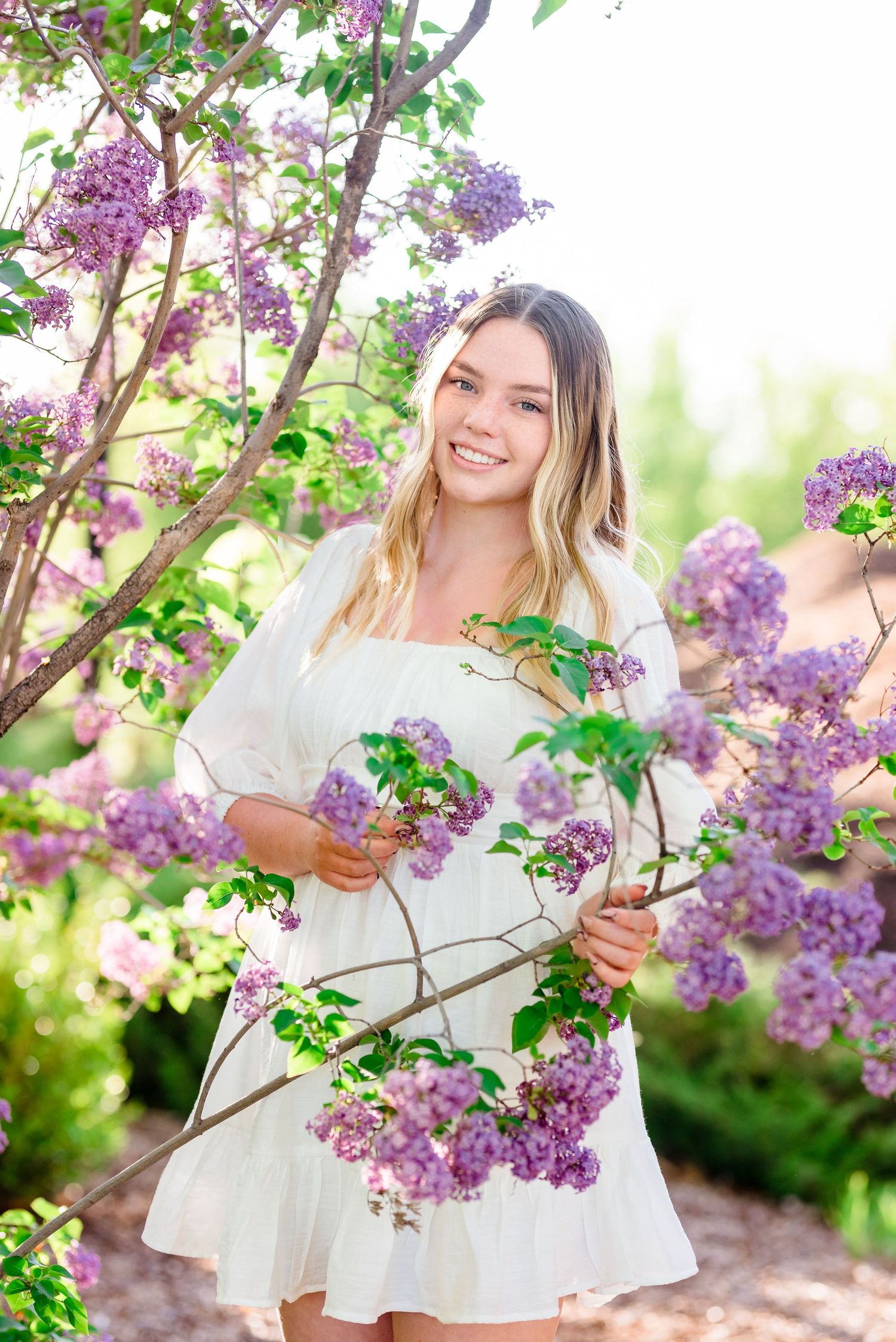 Spring portrait in a white dress among blooming purple lilac branches in soft natural lighting.