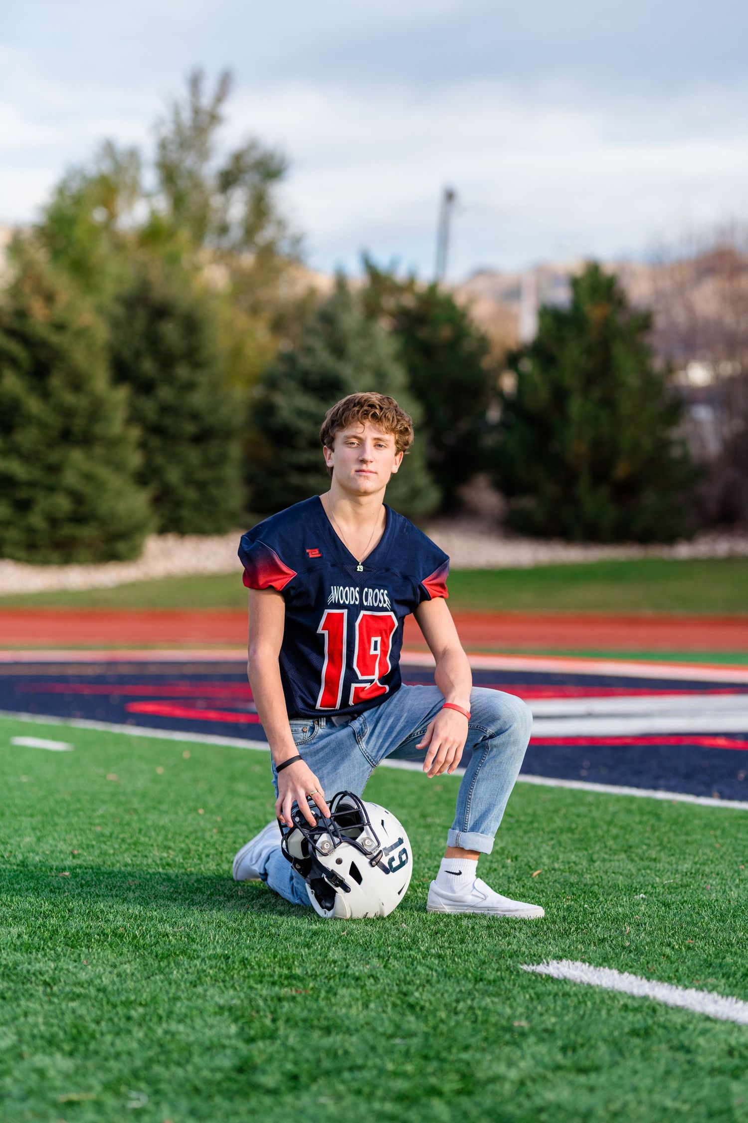 High school football player in navy jersey sits on field with helmet during outdoor sports portrait session.