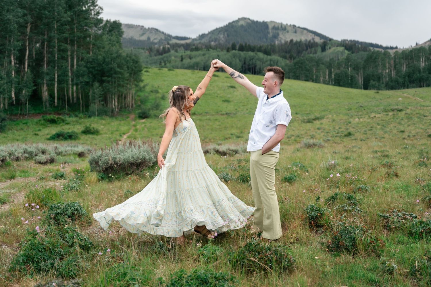A couple dances together in a scenic mountain meadow filled with wildflowers during a romantic outdoor photoshoot.
