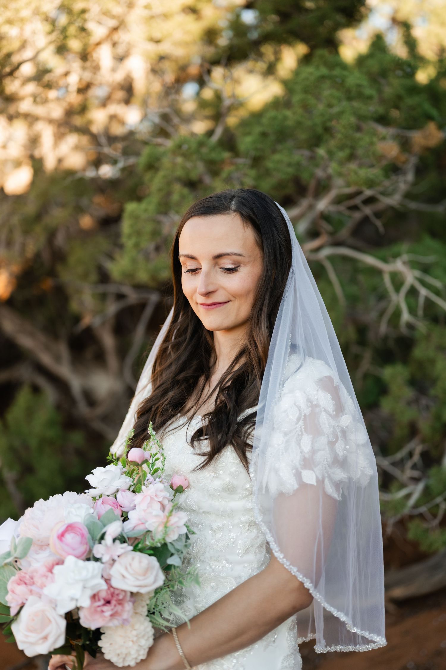 A bride holds a soft pink and white bouquet while wearing a delicate lace veil in natural lighting.