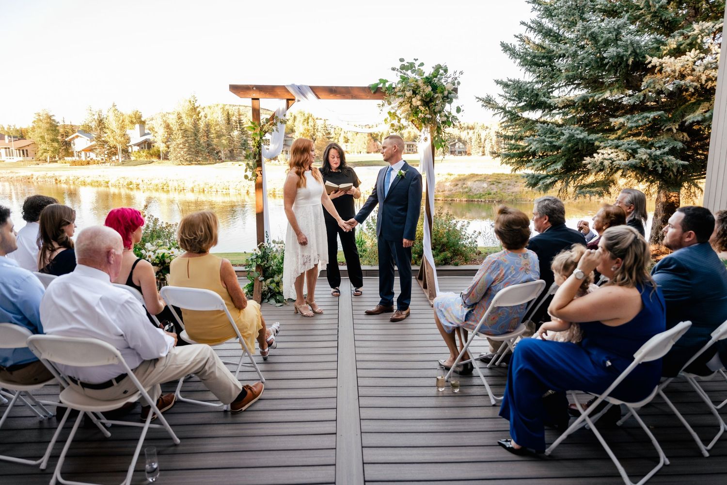 Outdoor wedding ceremony on a wooden deck with guests seated and evergreen trees in background.