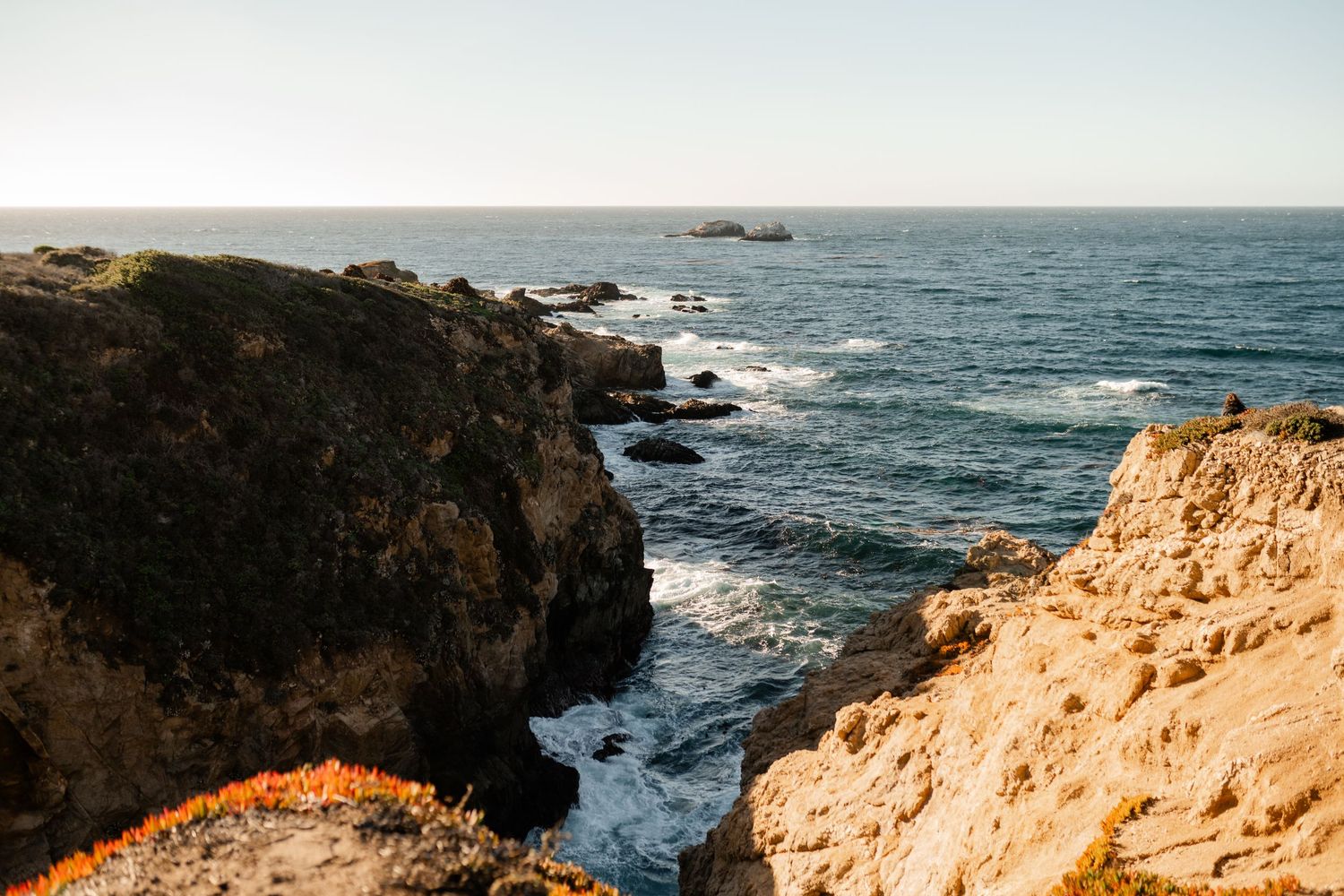 Rocky coastal cliffs and ocean waves under bright sky at sunset.