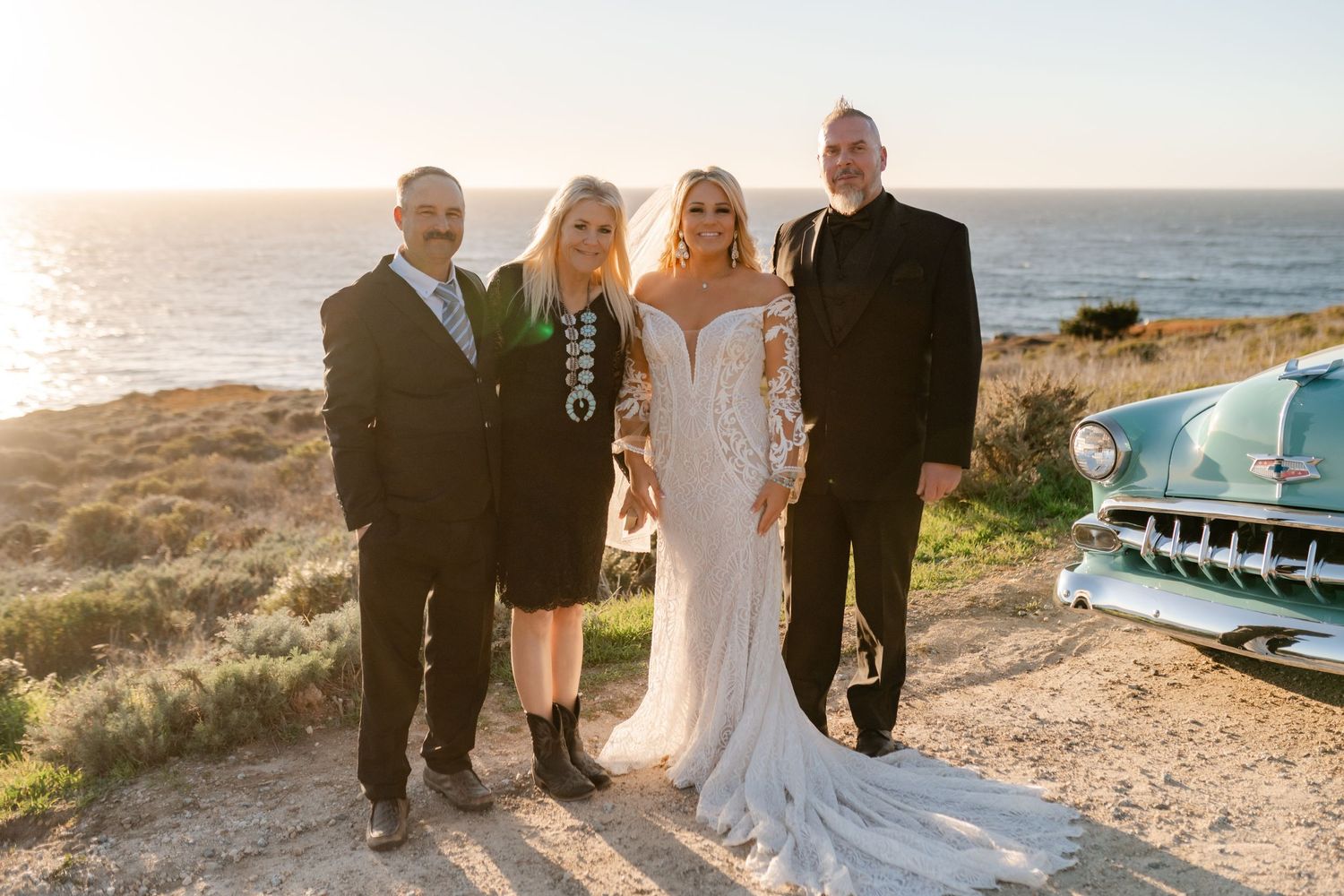 Wedding party posing by vintage car with dramatic coastal cliffs in background.