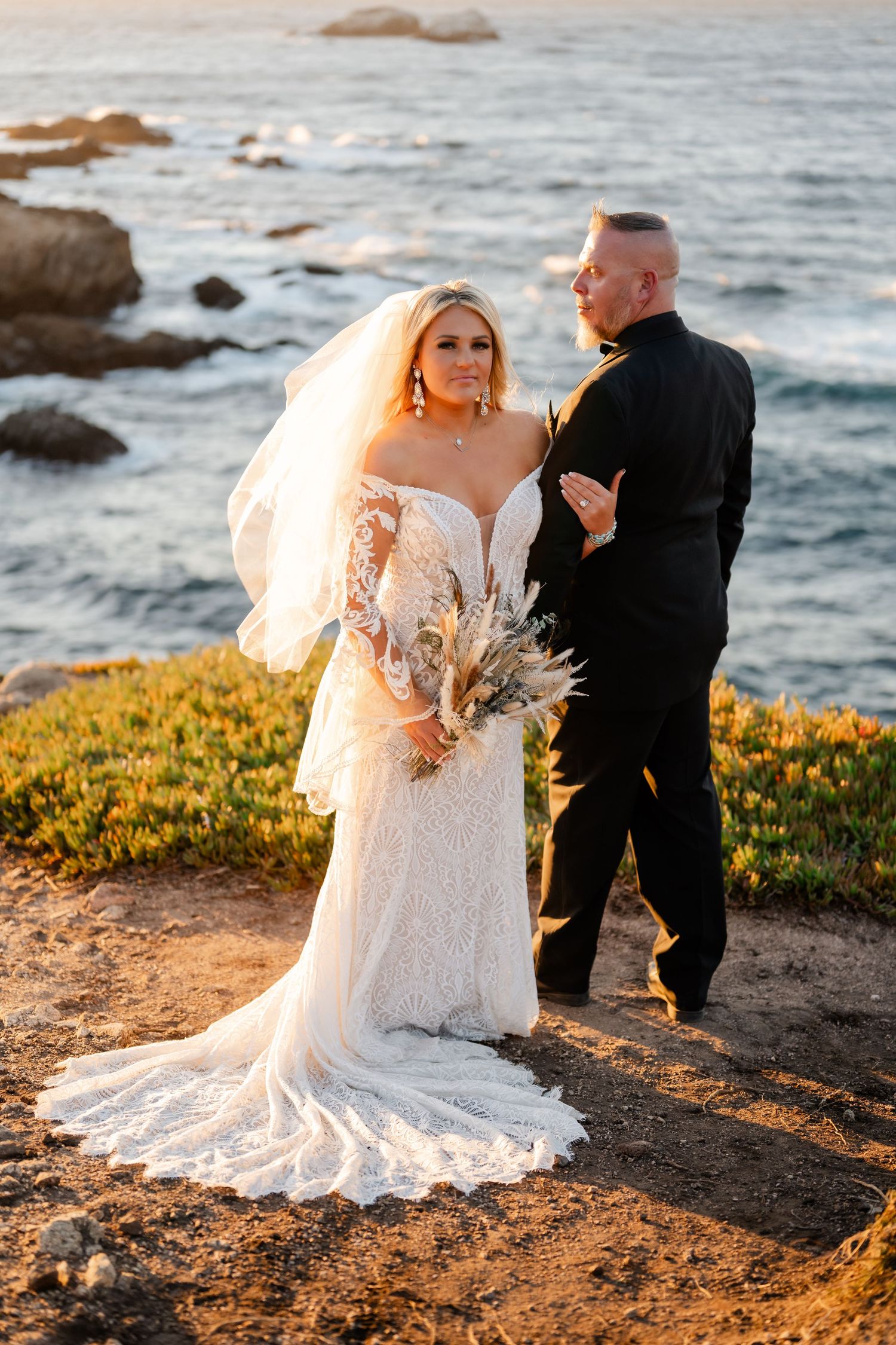 A couple poses on a coastal cliff with ocean waves and yellow wildflowers in the background.