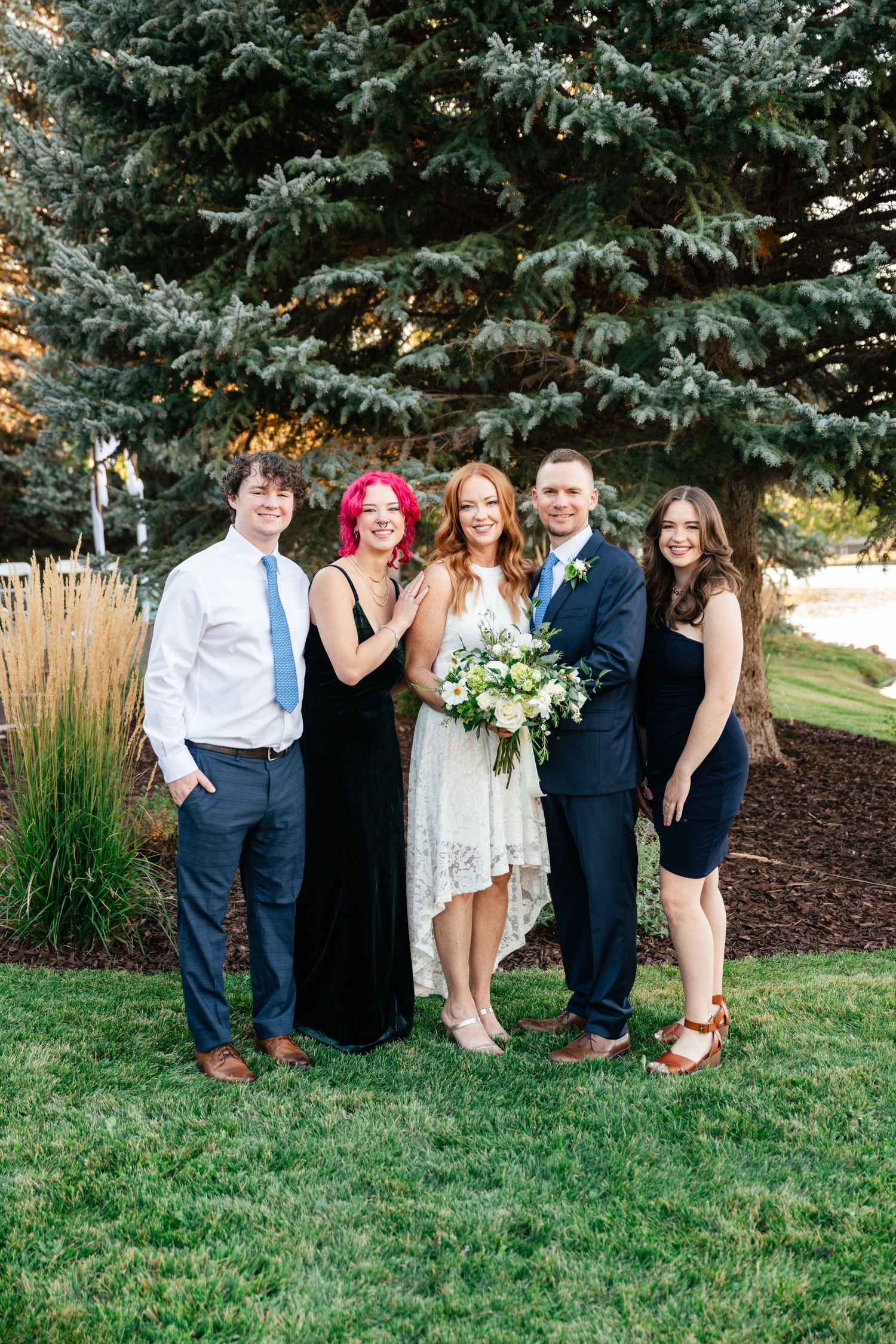 Wedding group photo outdoors with evergreen trees and ornamental grasses in the background.