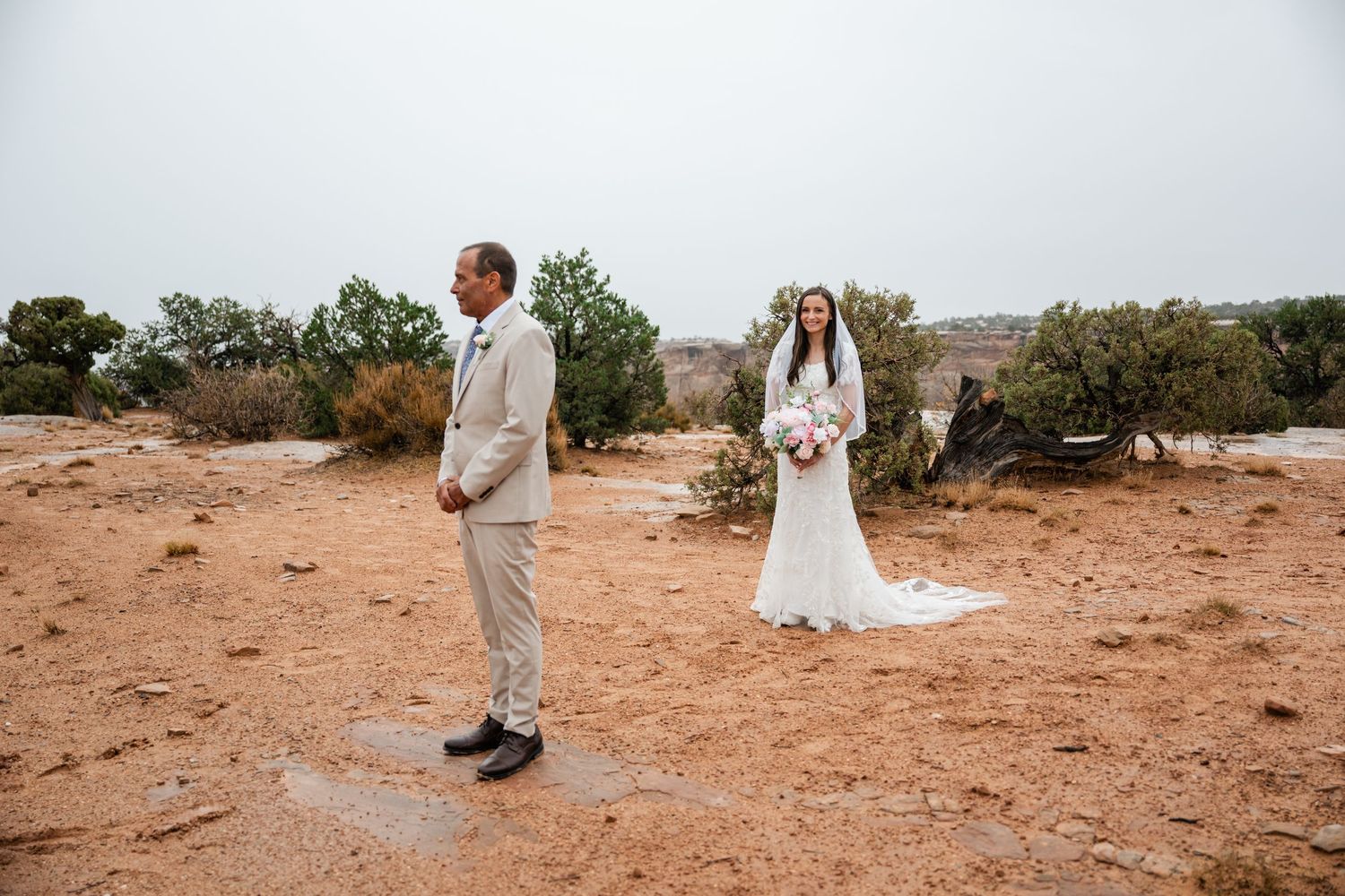 A couple in wedding attire stands together in a scenic desert landscape at sunset.