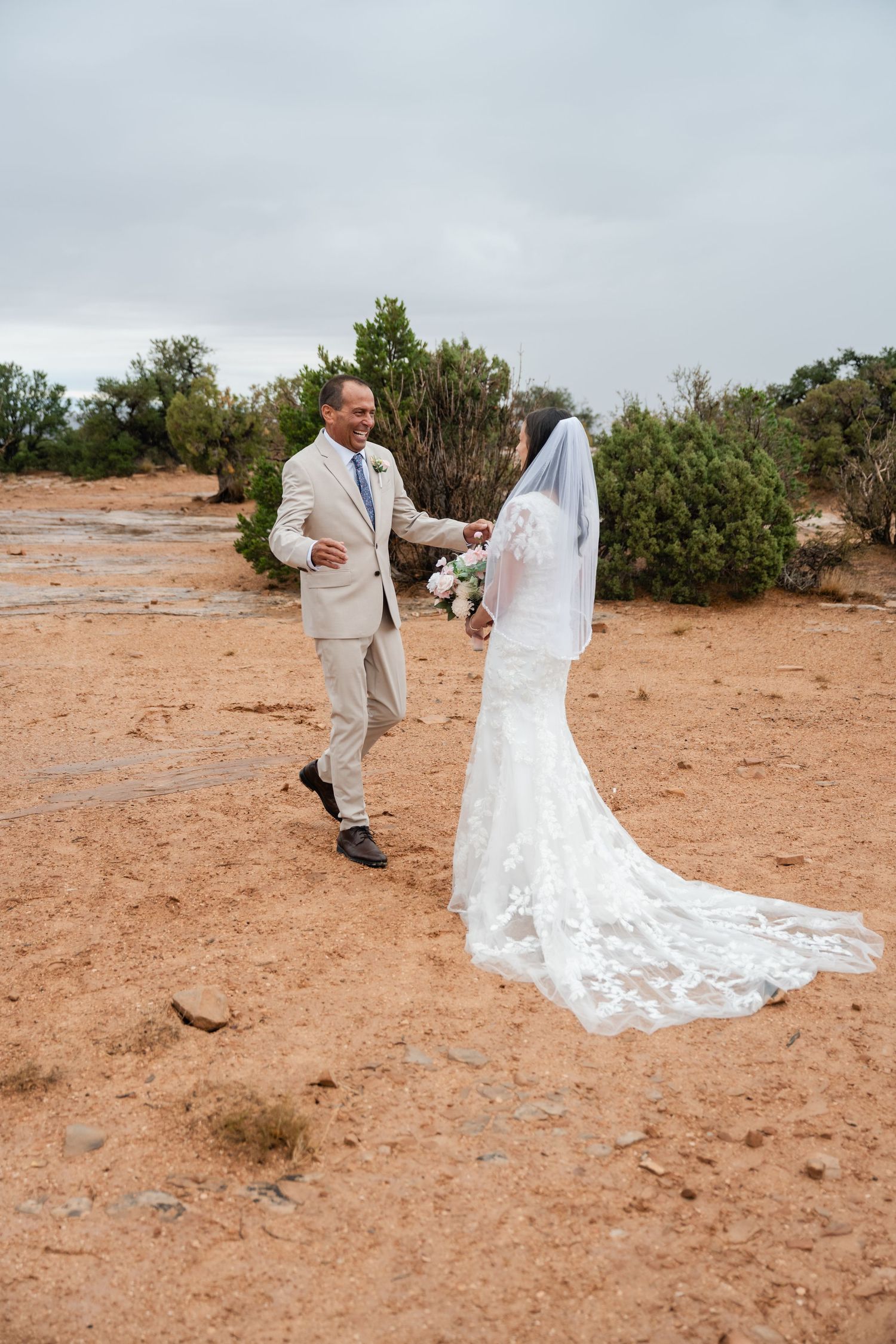 A couple walks on red desert terrain during their wedding photoshoot with the bride wearing a flowing white gown and veil.